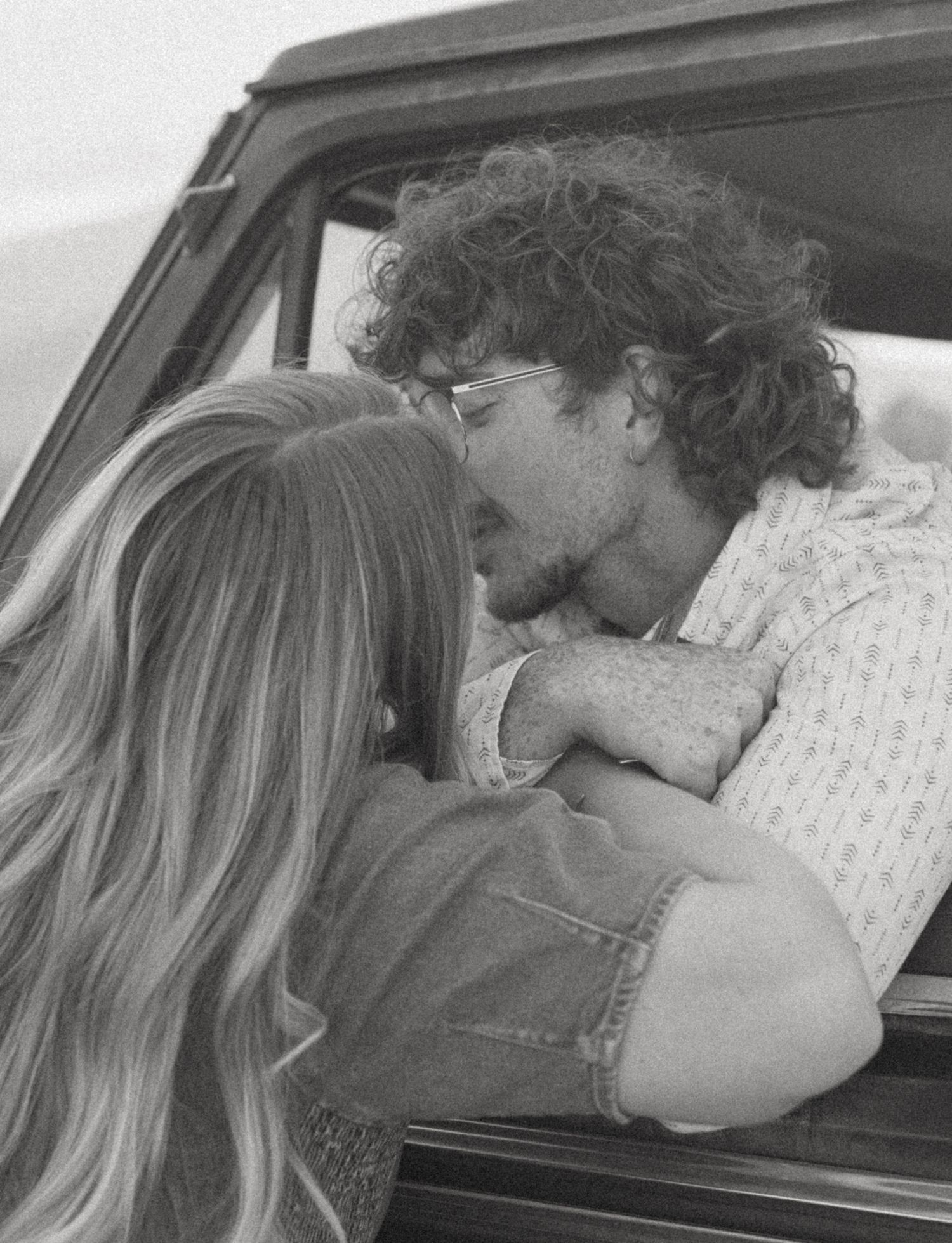 Black and white photo of curly-haired man leaning out of vintage truck window to kiss blonde woman tenderly.