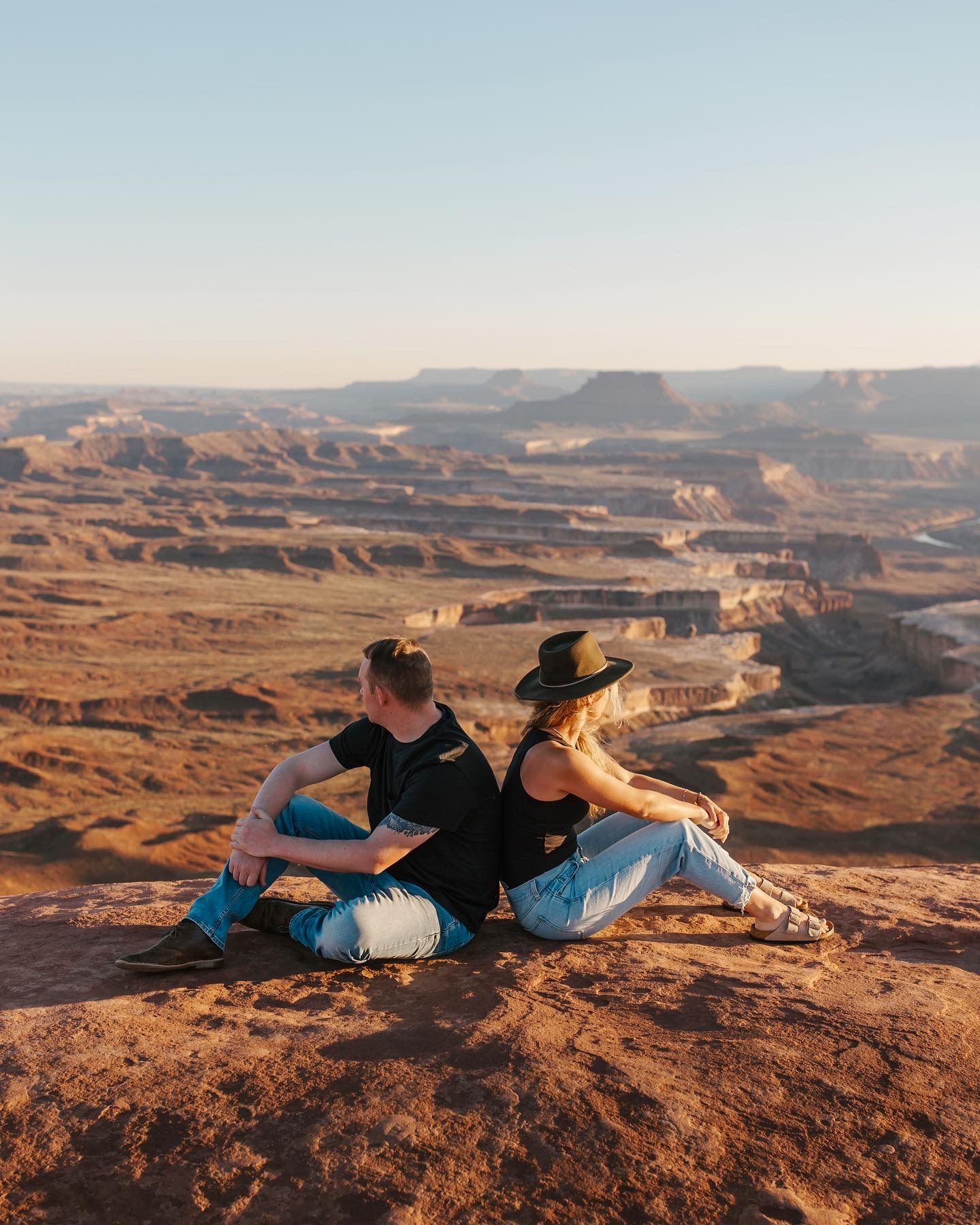 Couple sitting back to back on red rock cliff overlooking vast Canyonlands desert canyon at sunset.