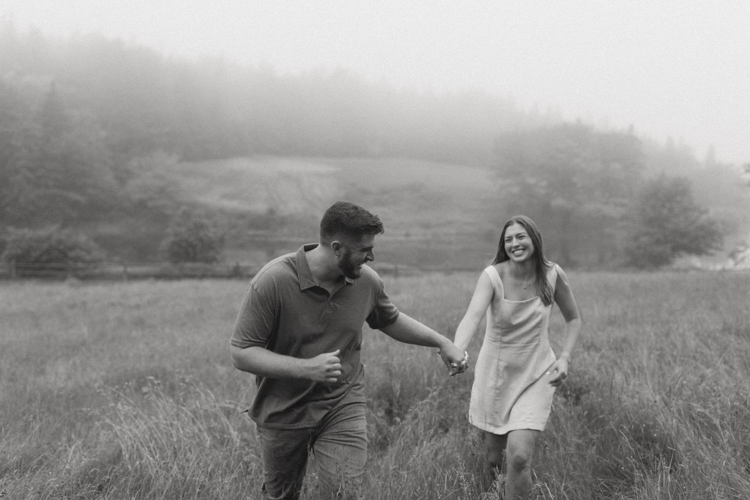 Black and white photo of a laughing couple holding hands while running through a misty meadow.
