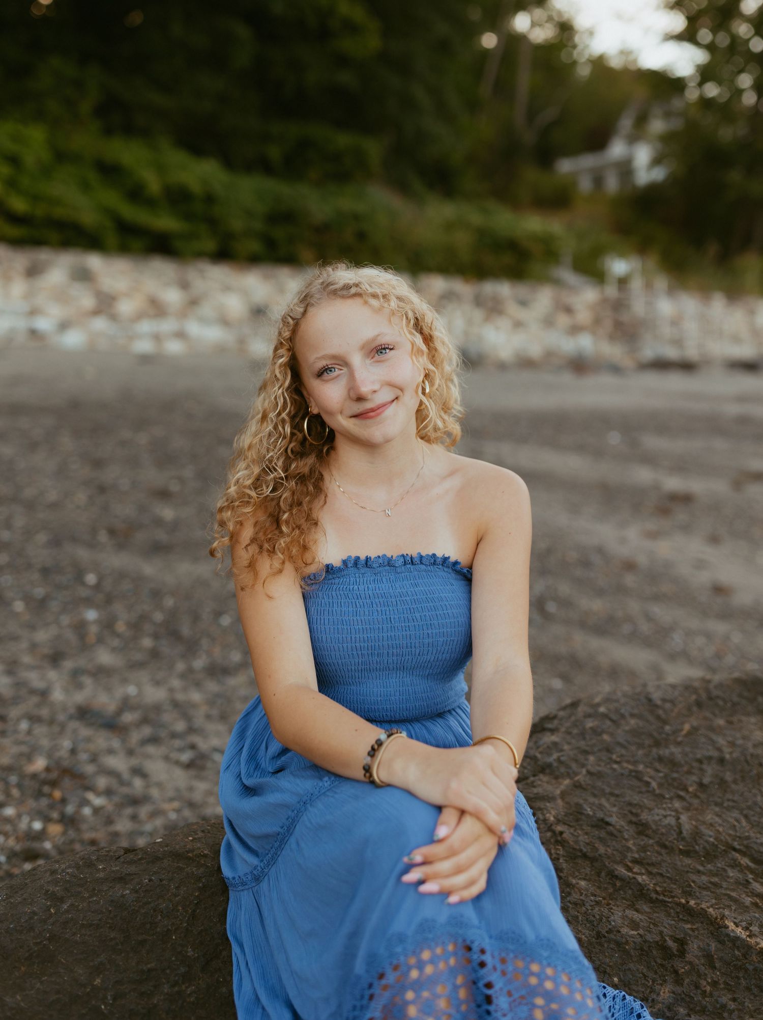 Young woman with curly blonde hair in a blue strapless dress sitting on a rock at a pebble beach.