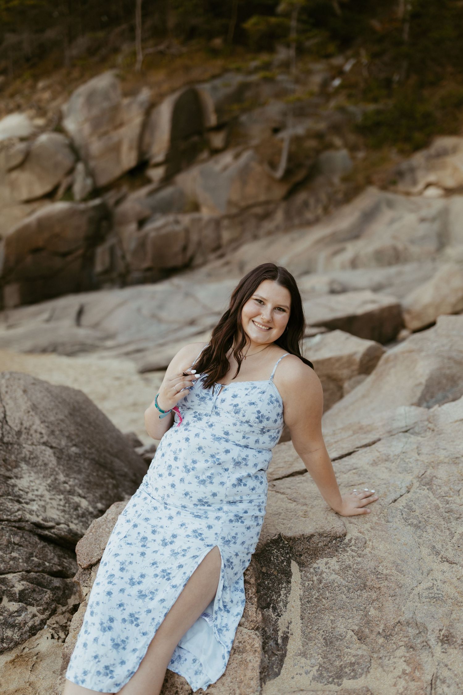 Woman in blue floral sundress posing on rocky coastal landscape, smiling at camera.