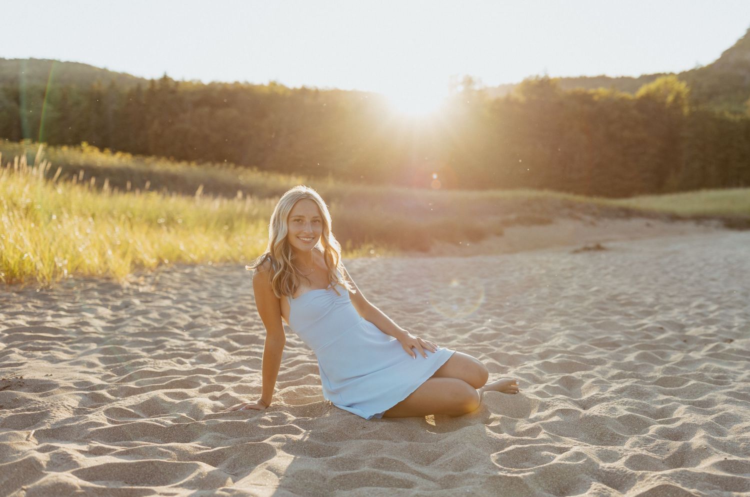 Young blonde woman in white dress sitting on sandy beach with golden sunset light behind her.