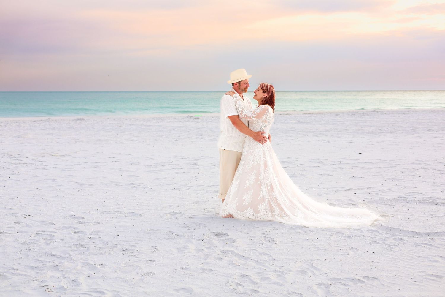 A couple in white wedding attire embraces on a sandy beach at sunset with turquoise ocean waves behind them.