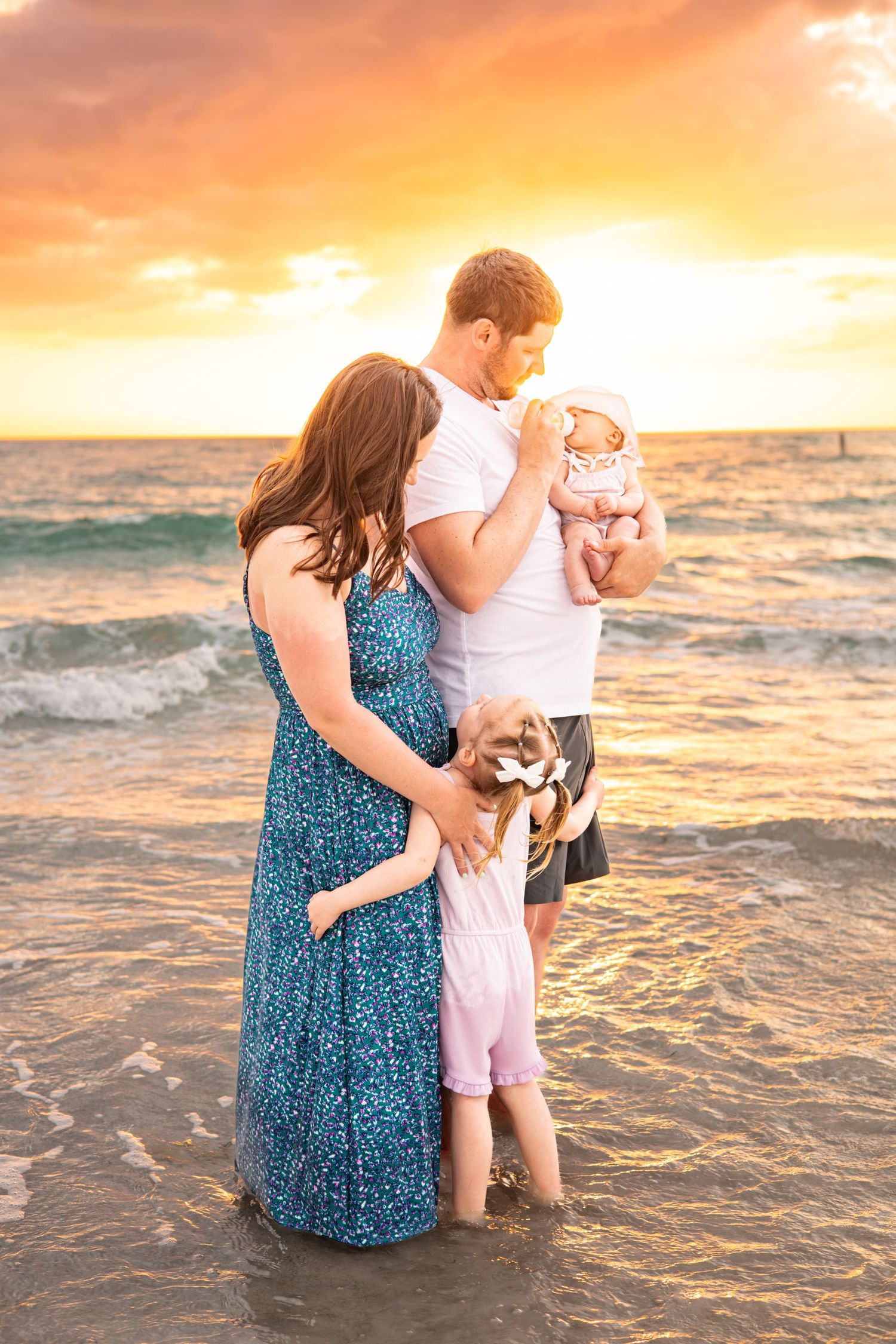 Family of four standing on a beach at sunset, parents holding newborn baby and toddler, waves in background.