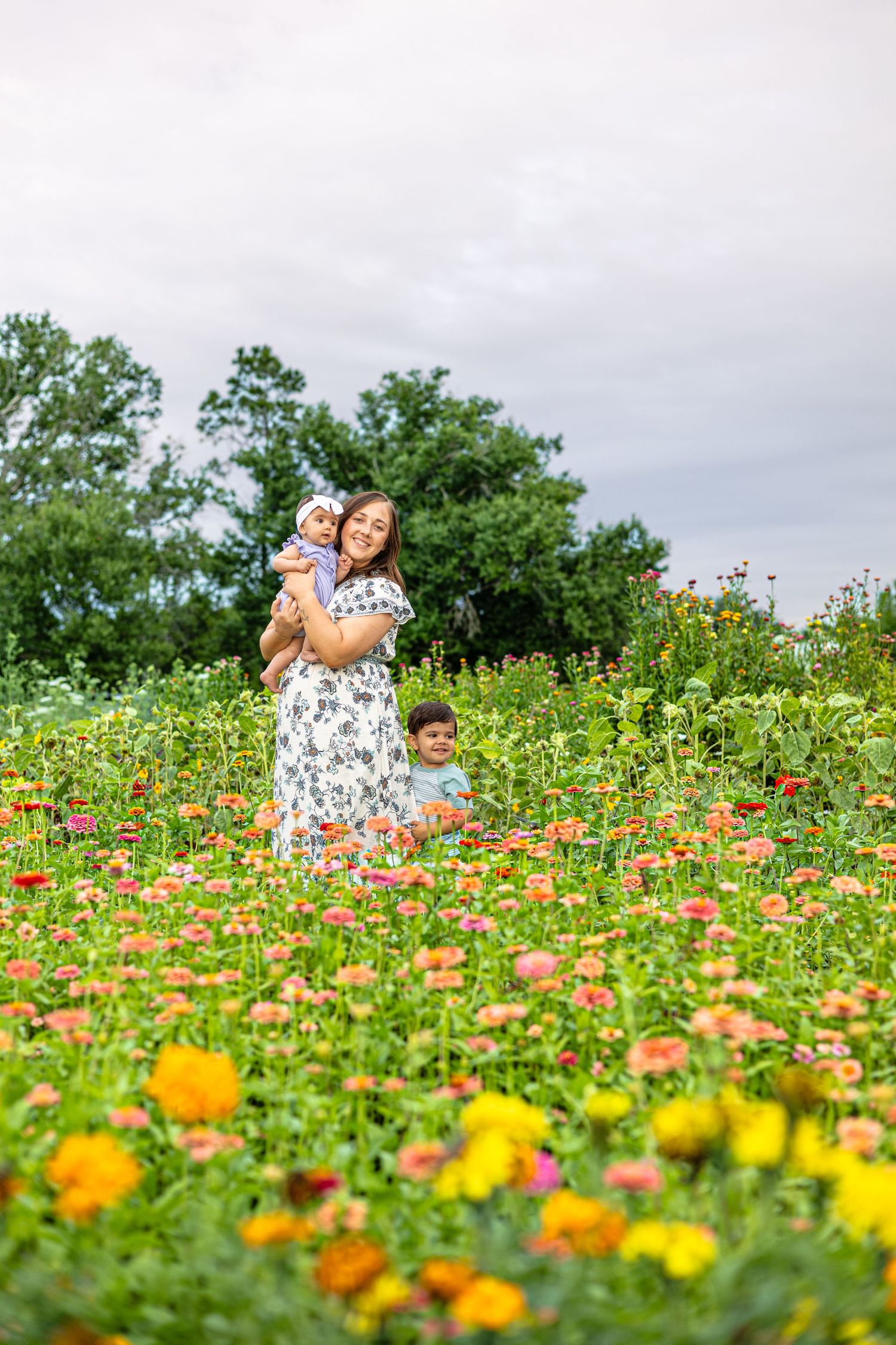 A smiling woman holds a baby in a colorful flower field with a child nearby, under a cloudy sky.