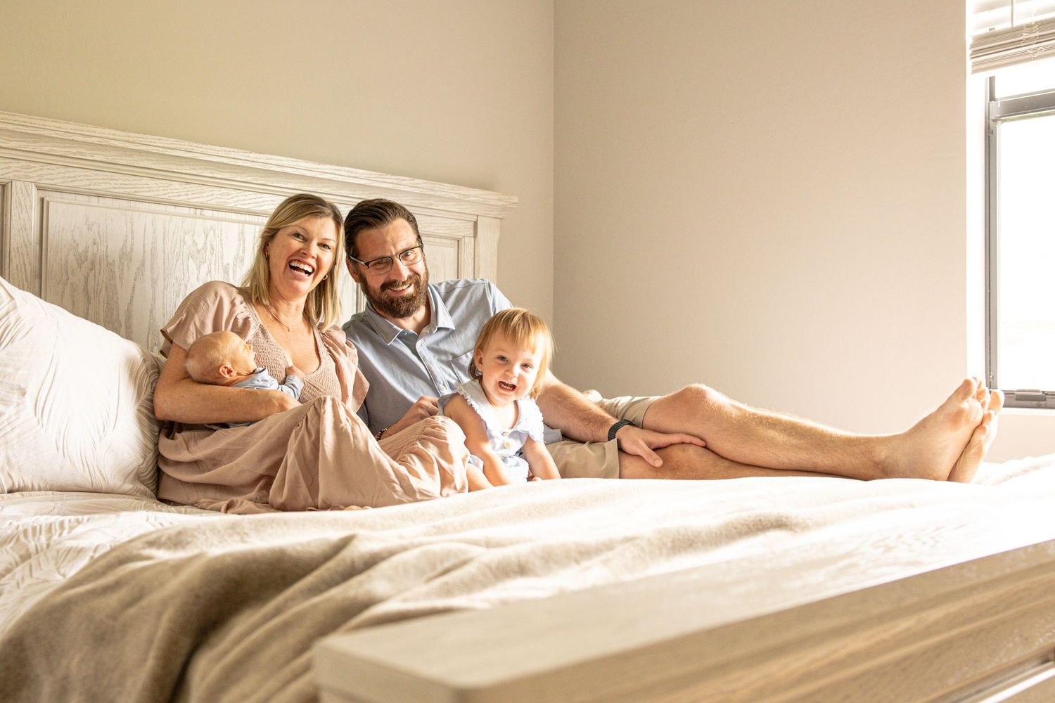 Happy family of four relaxing on a bed, parents smiling with newborn baby and toddler in bright bedroom.
