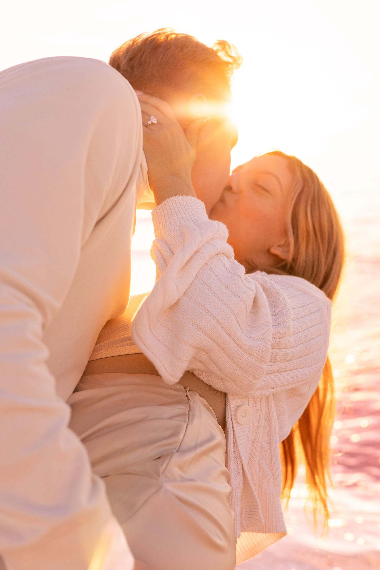 Couple sharing a romantic kiss at golden sunset by the water, both wearing cozy white sweaters.Sarasota Florida engagement photographer