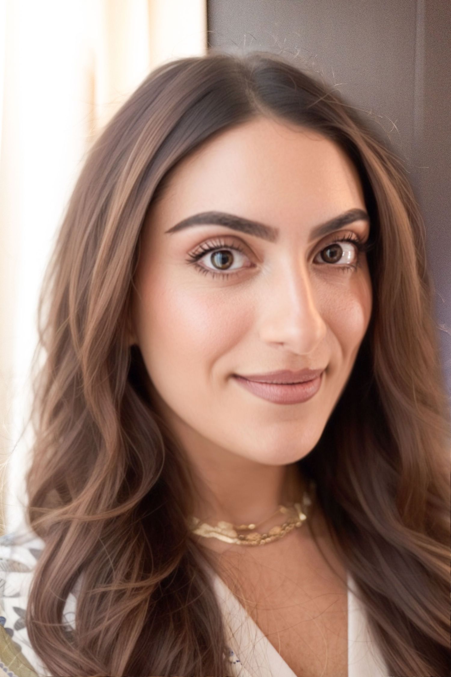 Young woman with long brown hair and gold necklace smiling softly in a bright, elegant setting.