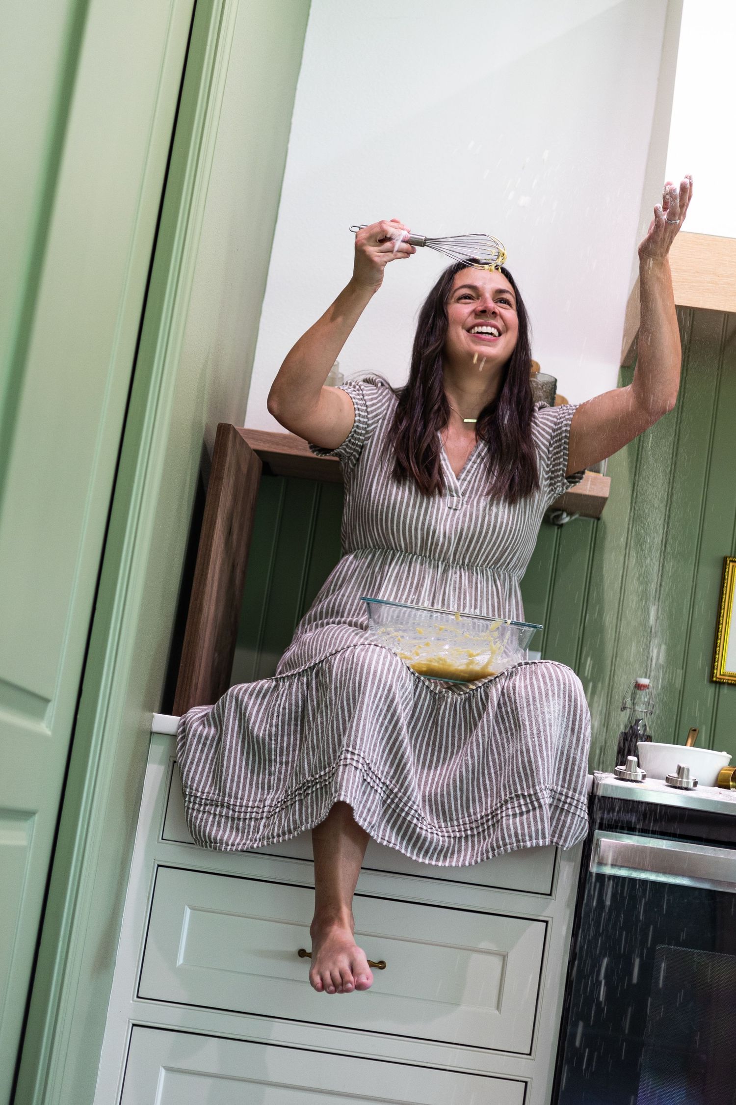 A joyful woman in a striped dress sits on kitchen counter, playfully tossing batter with a whisk over a mixing bowl.