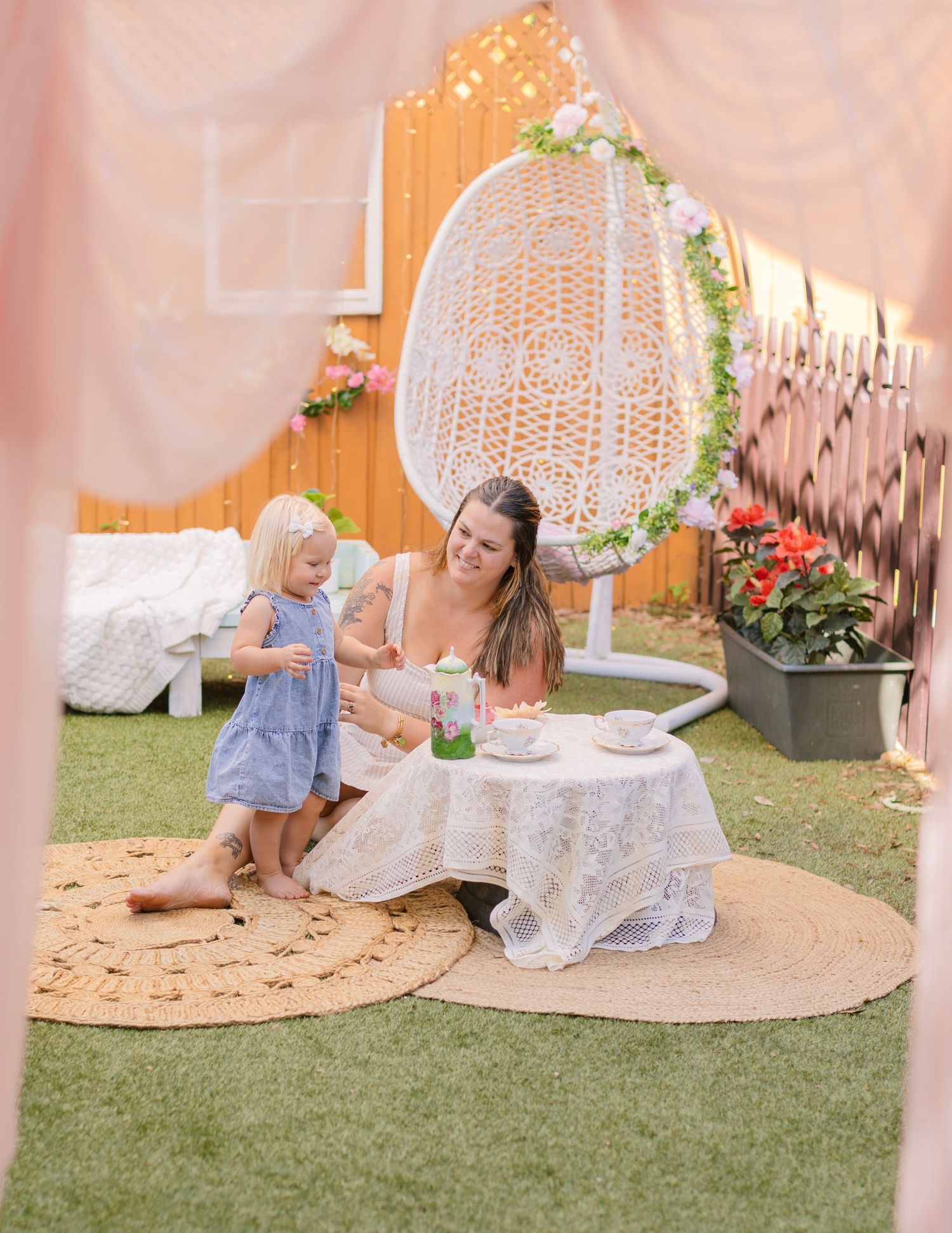 A woman and toddler girl enjoy a tea party on a jute rug in a decorated backyard with a hanging egg chair.