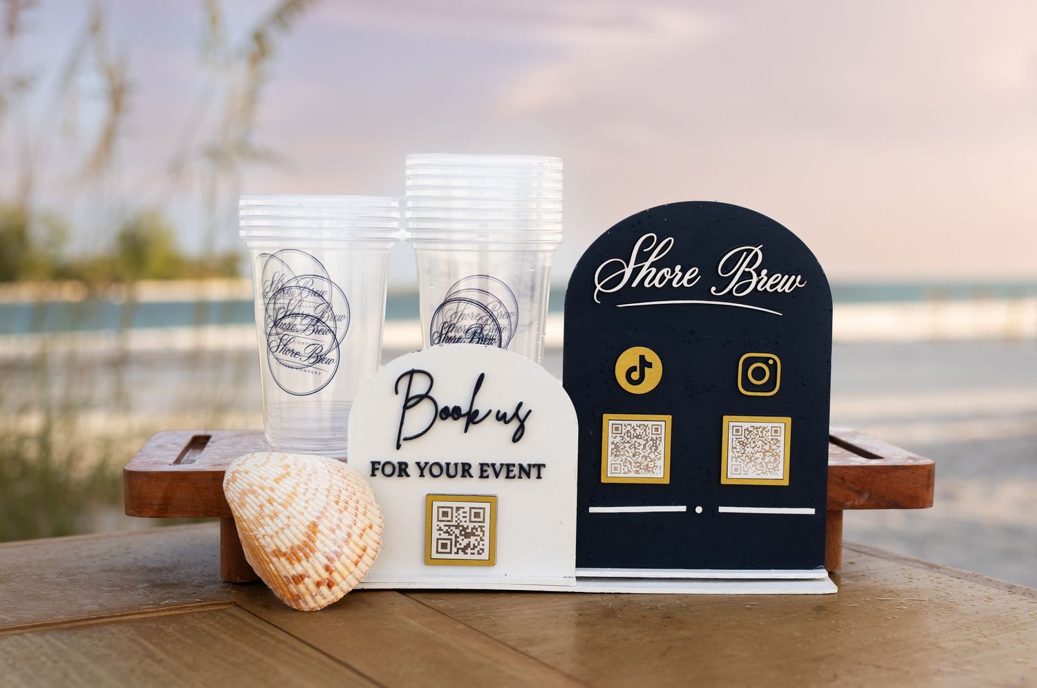 Shore Brew event booking display with QR codes, social media icons, branded cups, and a seashell on a wooden tray at the beach.
