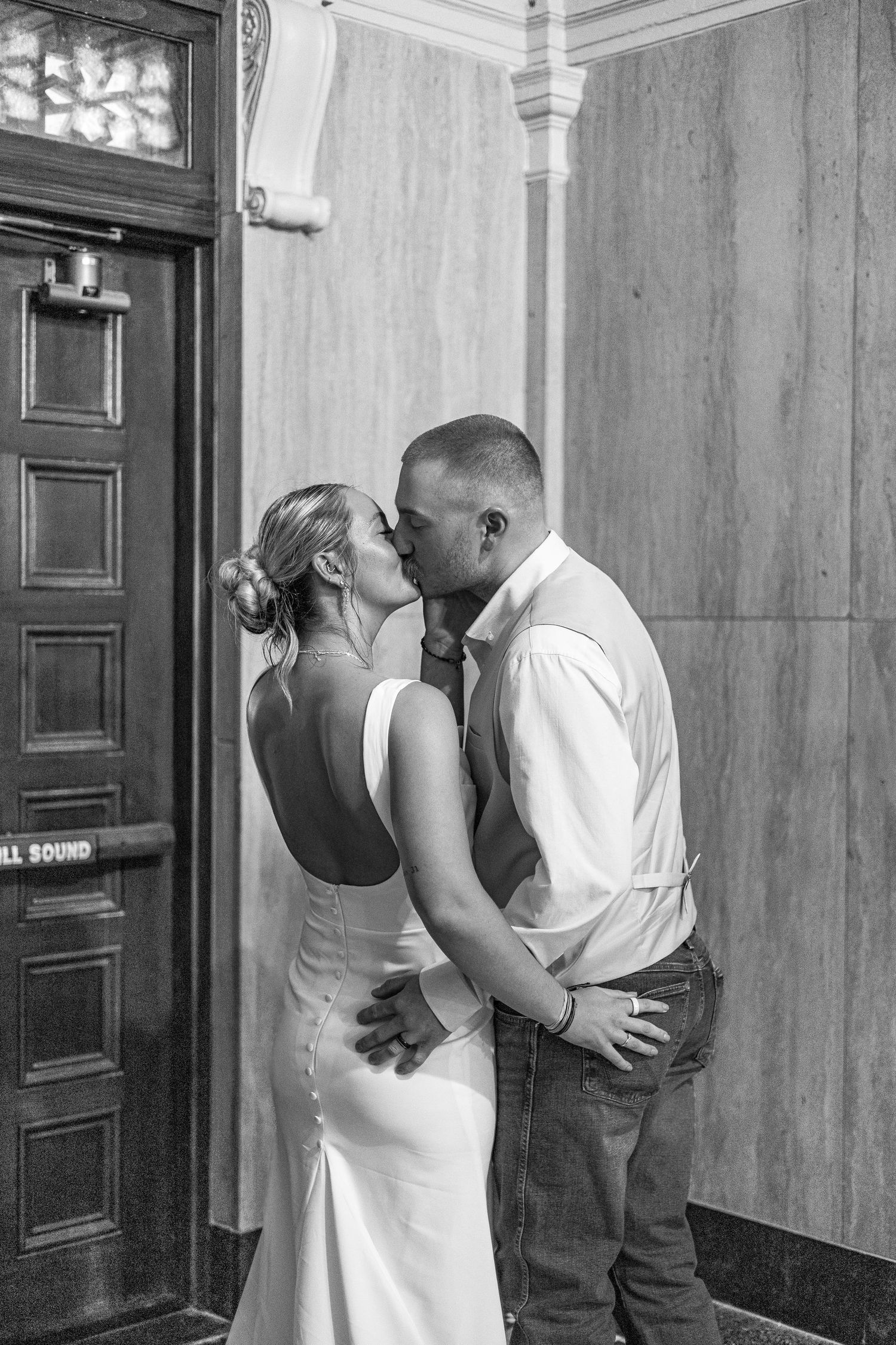 Black and white photo of a couple sharing a romantic kiss in wedding attire outside an elegant building entrance.