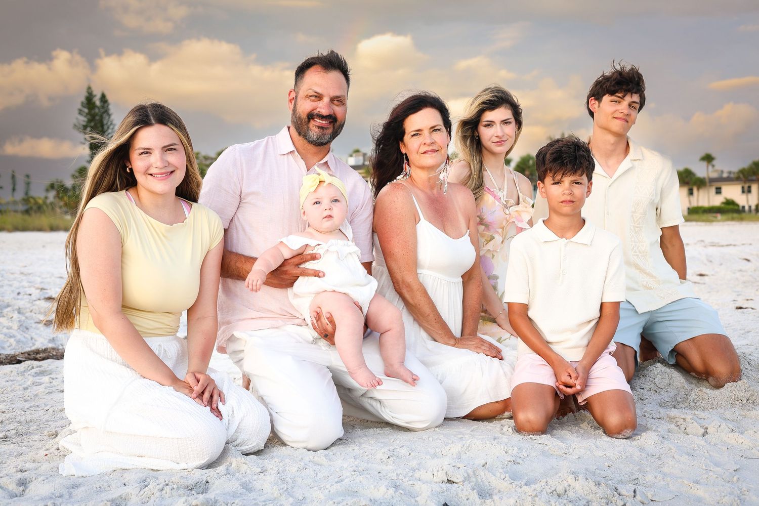 Multi-generational family of seven smiling together on a sandy beach at sunset in casual white and pastel outfits.