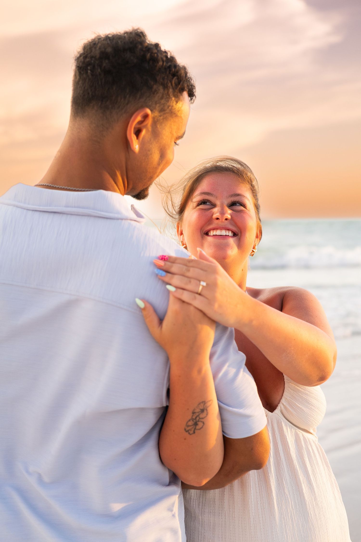 Couple embracing on beach at sunset, woman smiling wearing white dress and ring, man in white shirt by ocean.