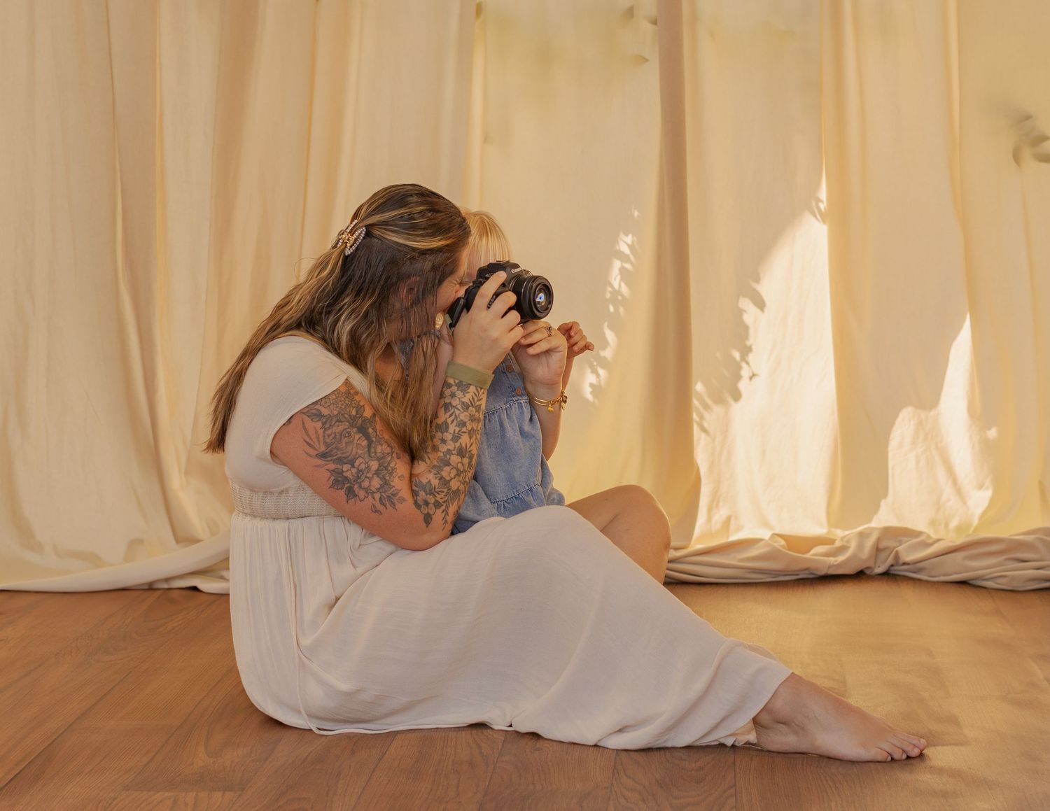 Tattooed woman in white dress sitting on wooden floor photographing with camera near flowing cream curtains.