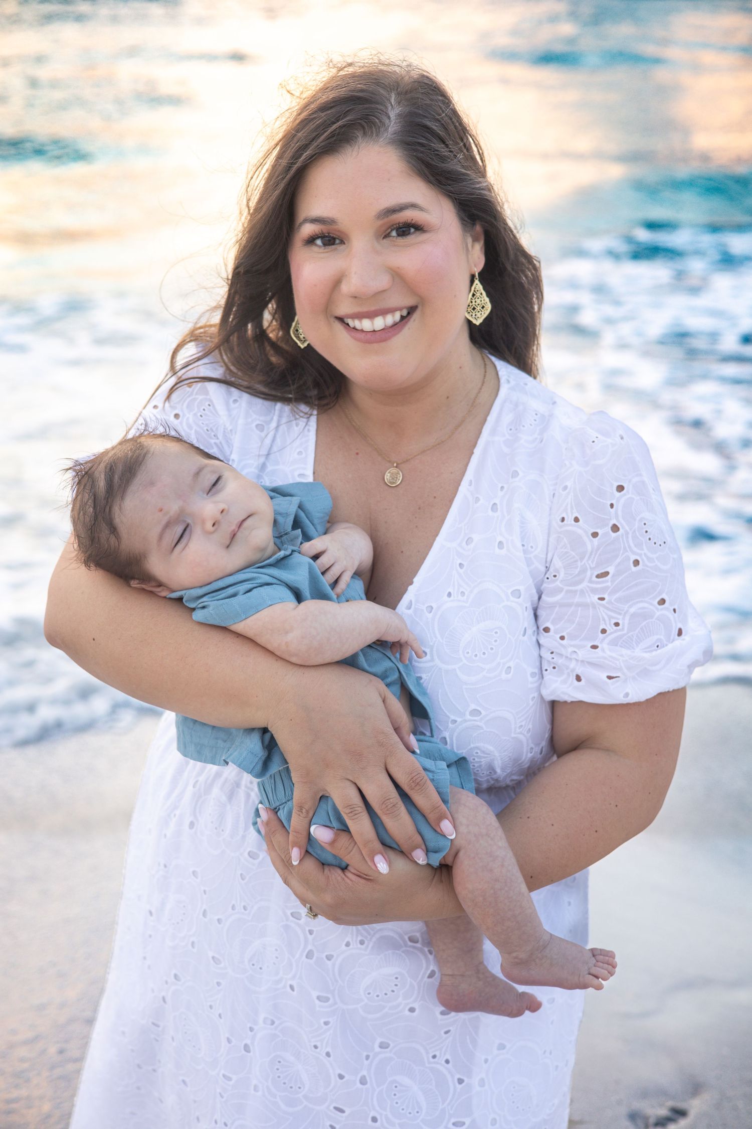 Smiling woman in white eyelet dress holds sleeping newborn baby in blue outfit on a sunny beach.