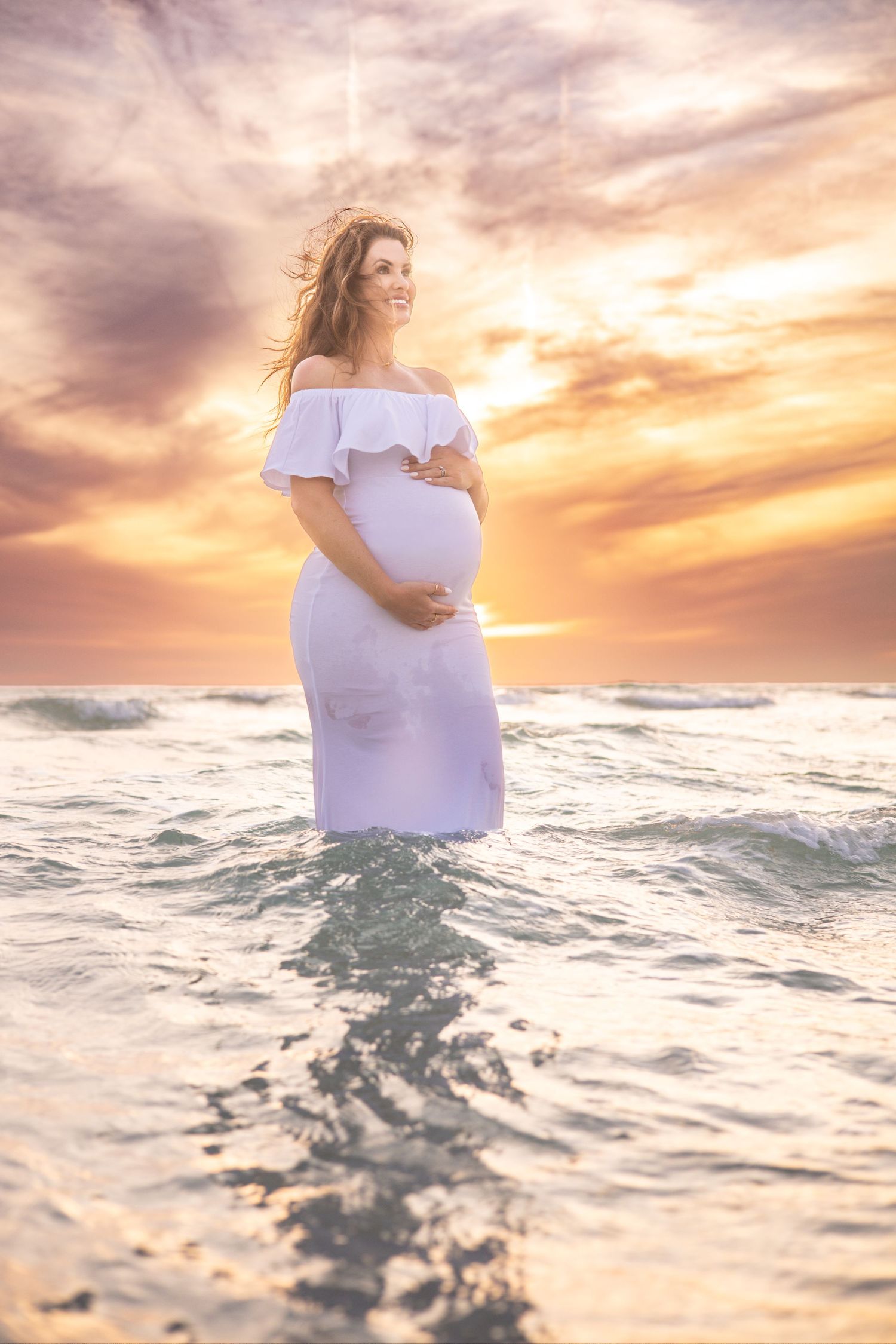 Pregnant woman in white off-shoulder dress standing in ocean waves at sunset, cradling her baby bump.