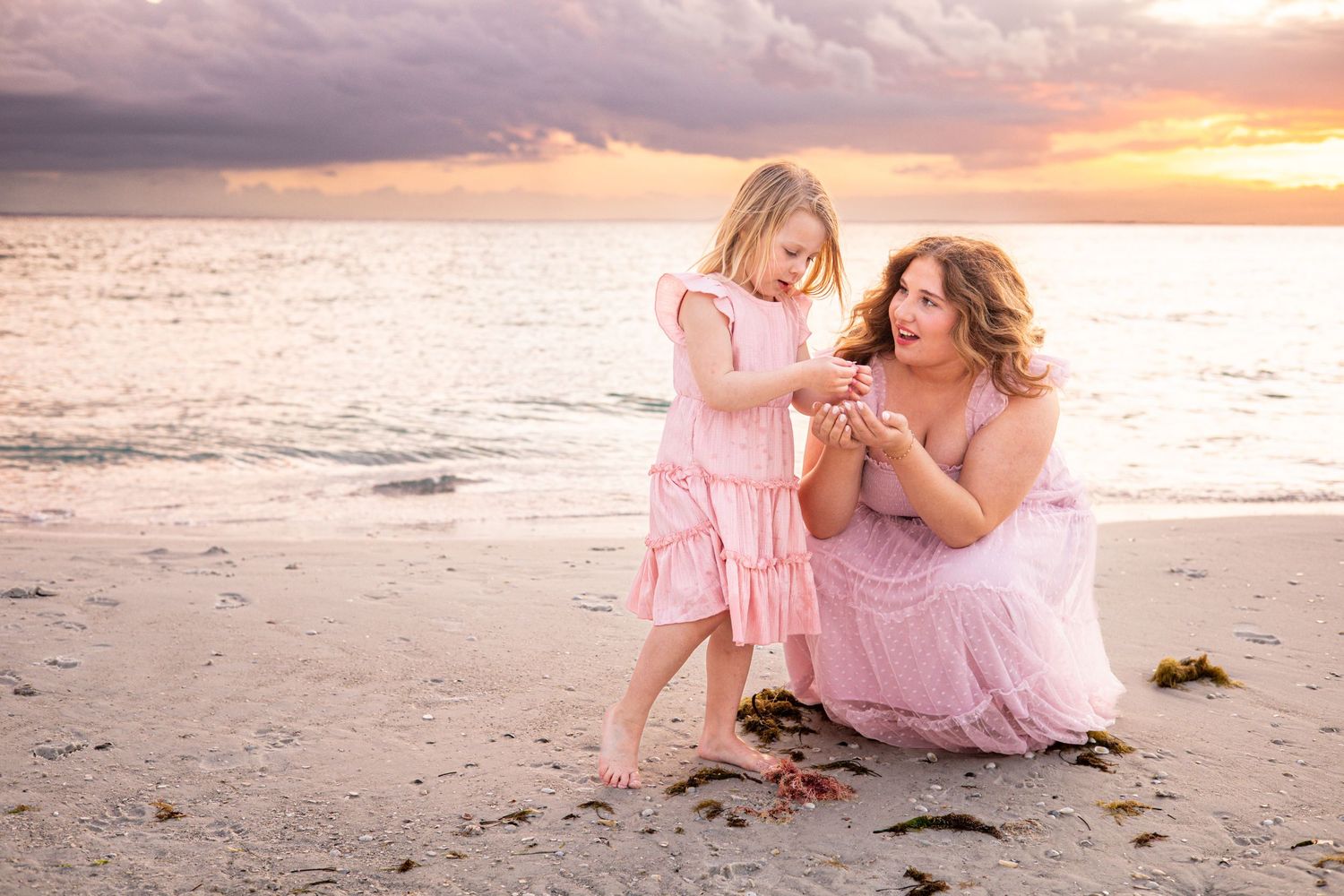 Mother and daughter in pink dresses share a tender moment on a sandy beach at sunset.