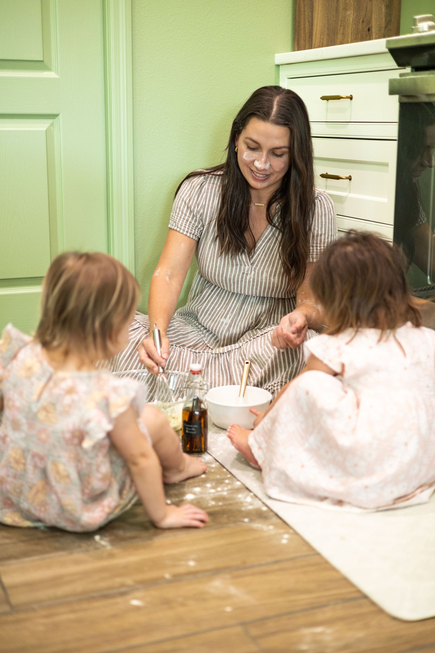 A smiling mother sits on kitchen floor baking with two young daughters, mixing ingredients in bowls together.