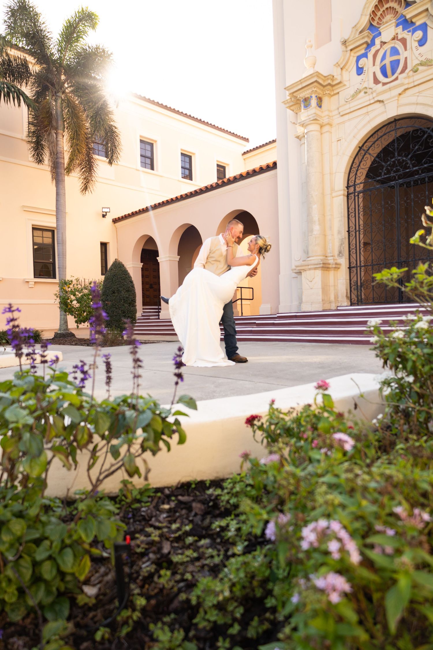 Bride and groom share a romantic dip kiss outside a Spanish Colonial Revival building surrounded by colorful flowers.
