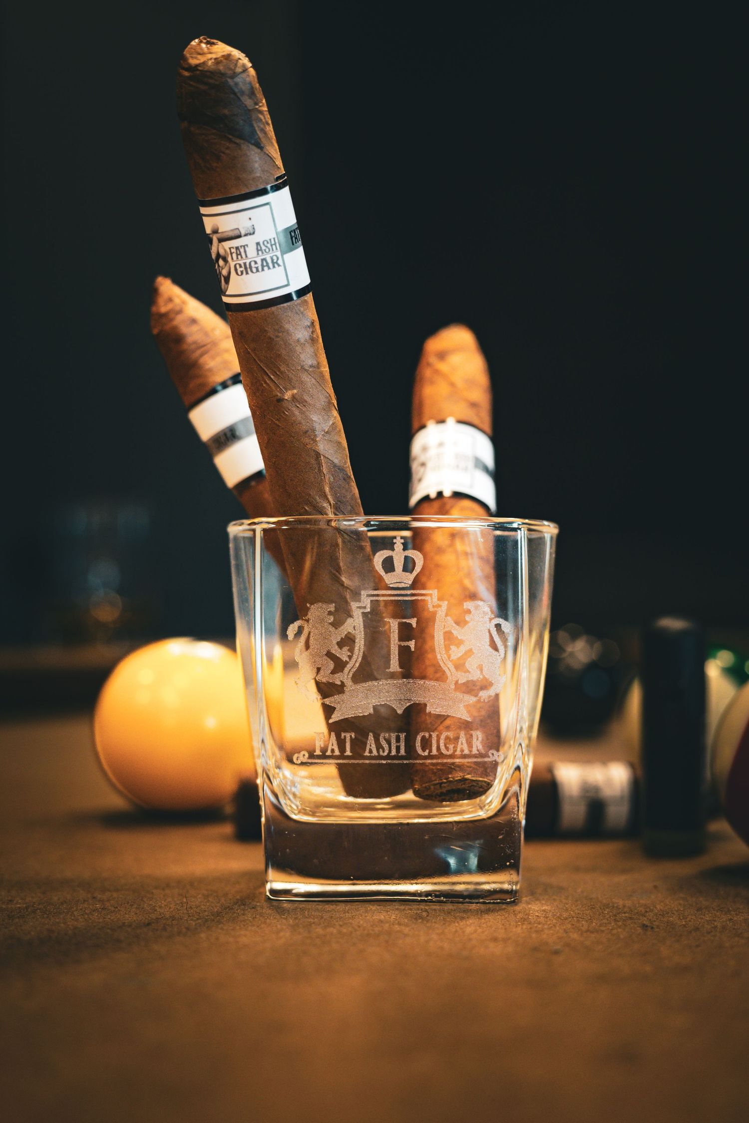 Fat Ash Cigar branded whiskey glass holding premium cigars on a rustic surface with moody dark background.
