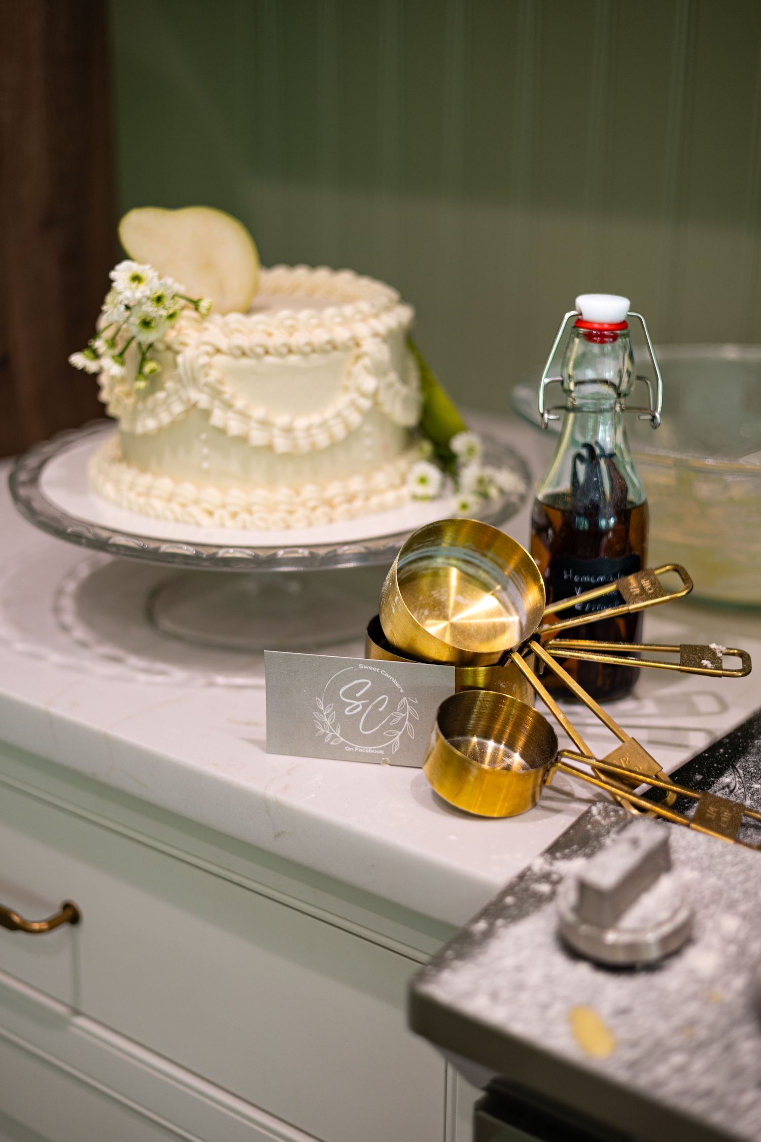 White wedding cake on stand beside gold measuring cups and vintage bottle on white kitchen counter.
