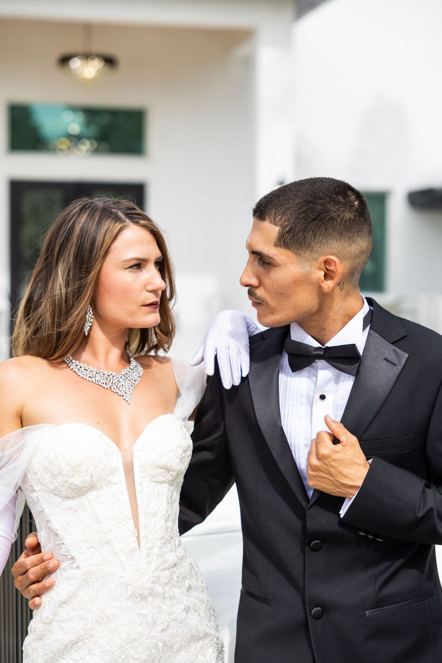 Elegant bride in white lace gown with diamond necklace and groom in black tuxedo sharing an intimate moment outdoors.