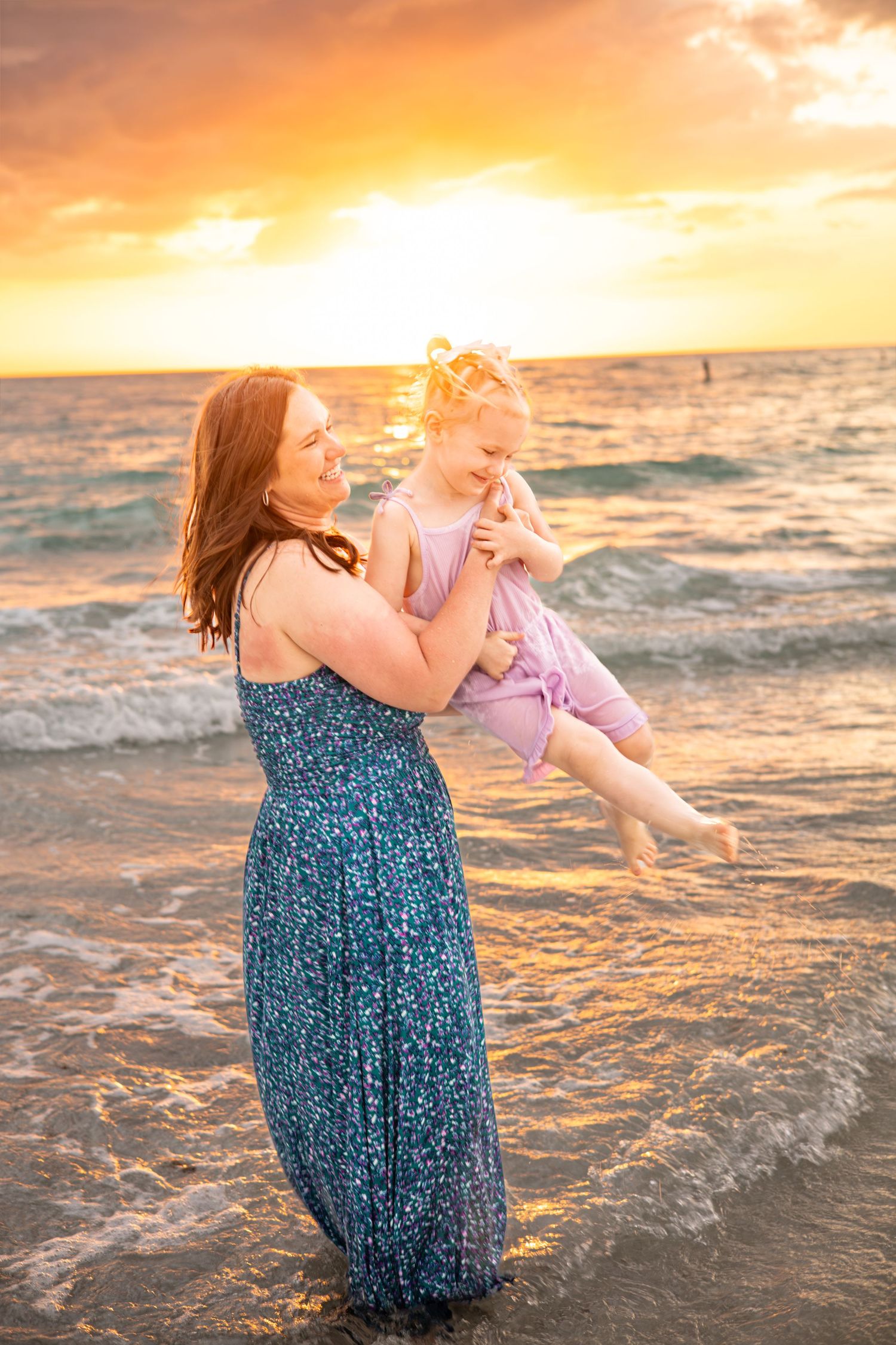 Mother holding young daughter on beach at golden sunset, waves lapping shore, woman in blue floral dress, child in pink outfit.