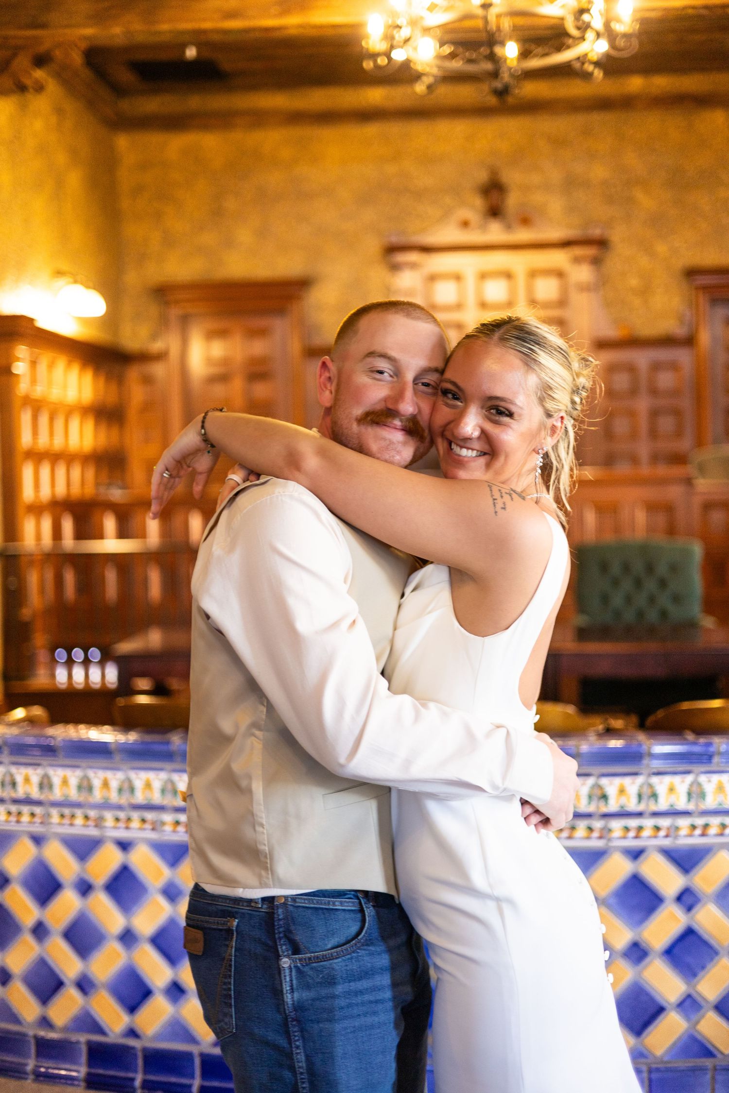 A smiling couple in white wedding attire embraces in a warmly lit venue with blue and yellow tiled walls.