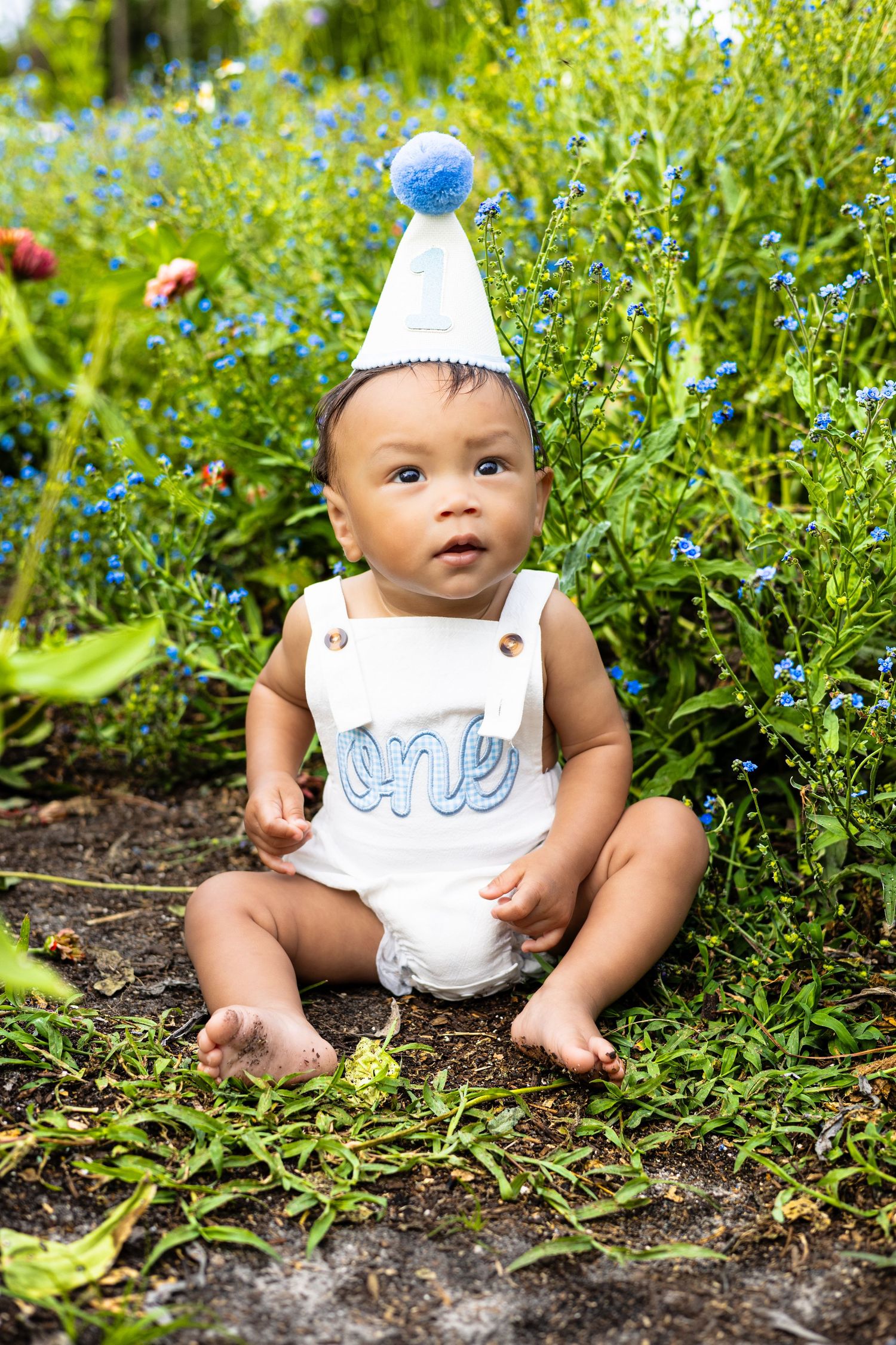 Adorable baby wearing a blue party hat and 'one' romper sitting outdoors among green plants for first birthday.
