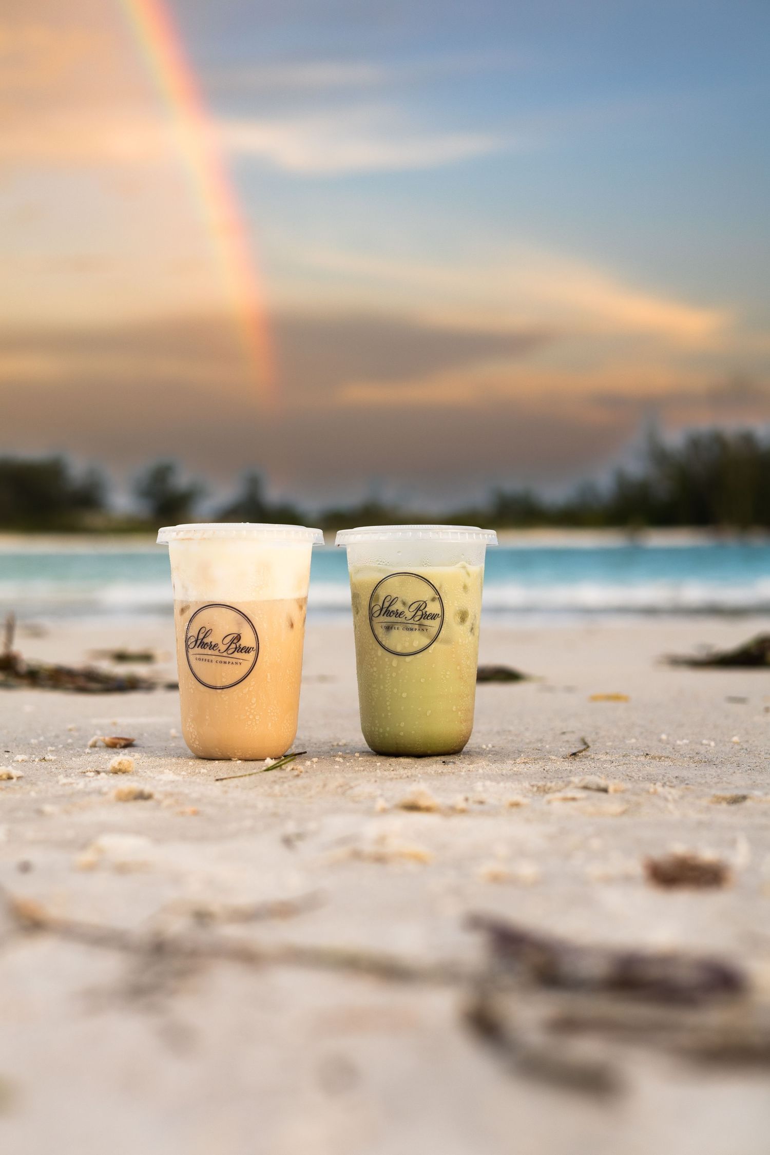 Two iced drinks on a sandy beach with a rainbow and ocean sunset in the background.