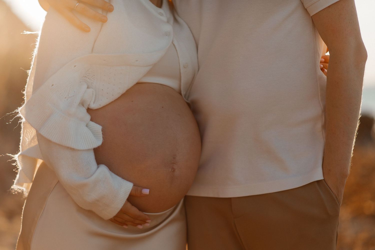 Expectant mother in a white top cradles her bare pregnant belly as her partner stands close beside her in warm golden sunset light.