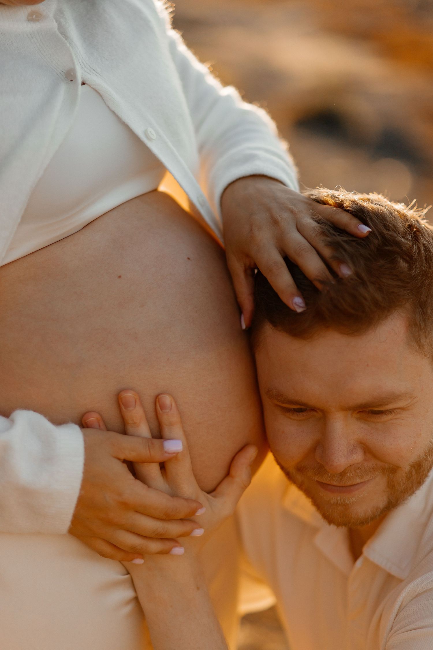 Man with a beard rests his ear on a pregnant woman's bare belly during a warm golden-hour maternity photoshoot.