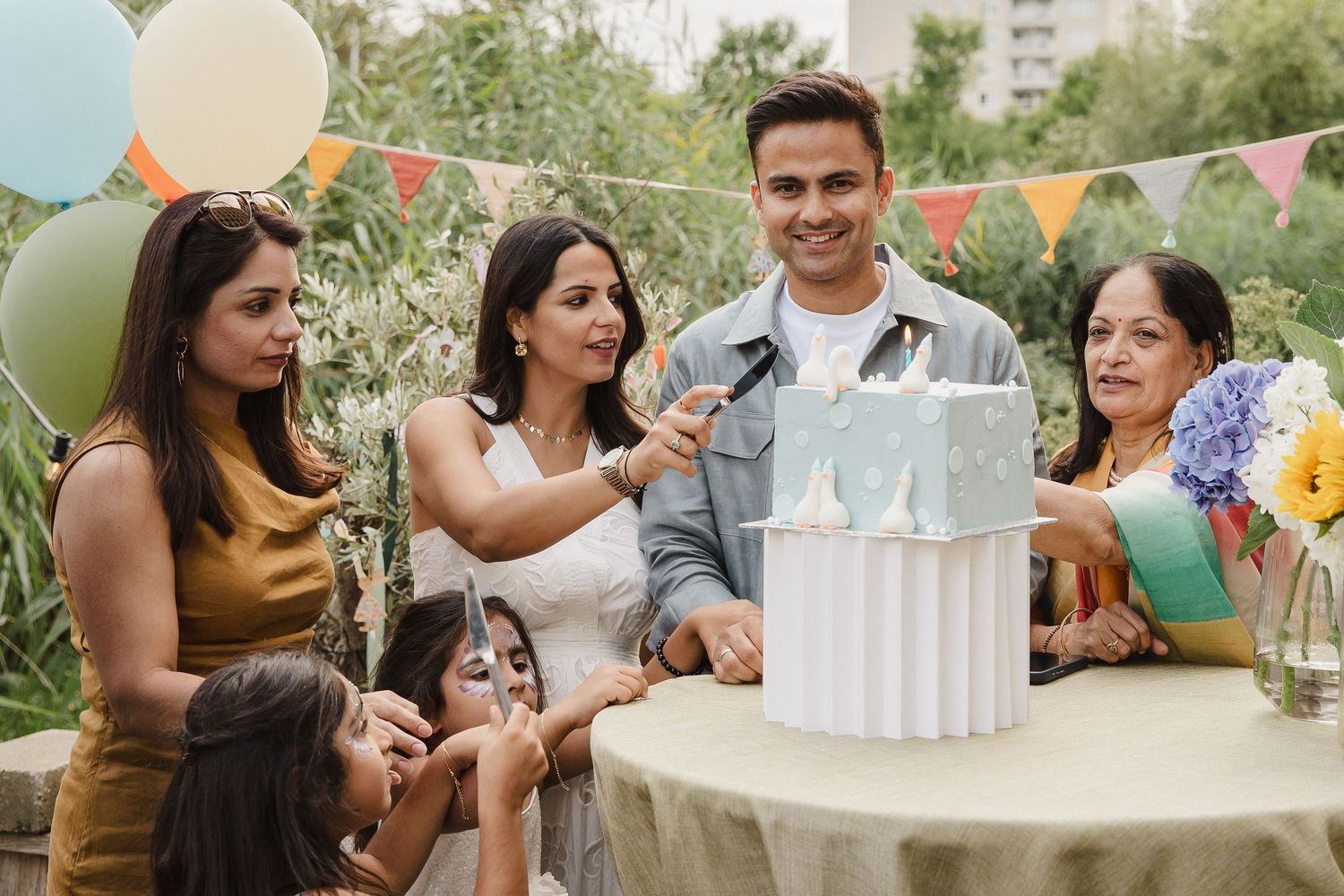 Smiling family and friends gather around a table to celebrate with a decorated cake at an outdoor birthday party with balloons and bunting.