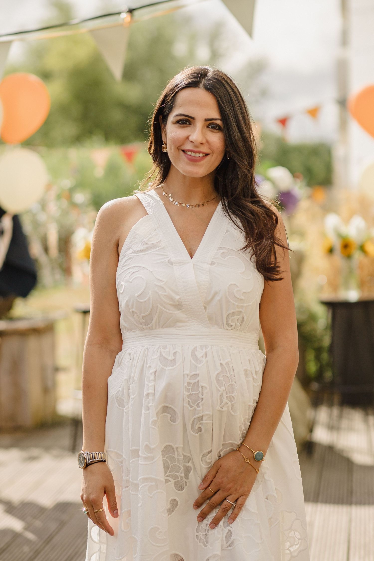 Smiling woman with long dark hair in a white lace sleeveless dress standing outdoors at a festive garden party with balloons and bunting.