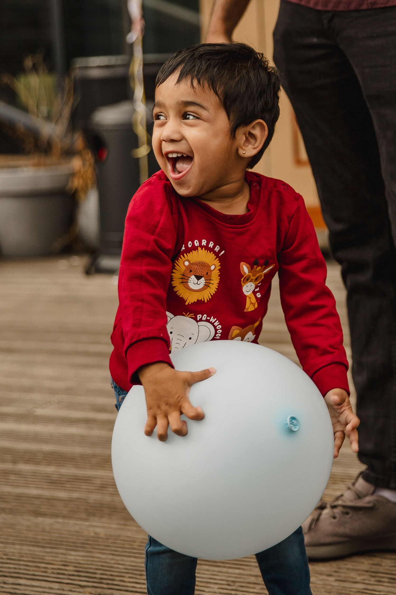 Smiling toddler in a red animal-print sweatshirt laughs while holding a large light blue balloon on a wooden deck.