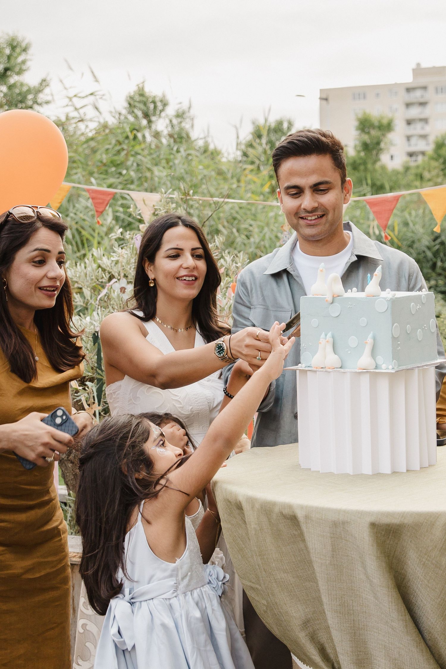 Smiling family gathers around a blue and white tiered cake at an outdoor birthday party with orange balloons and colorful bunting.