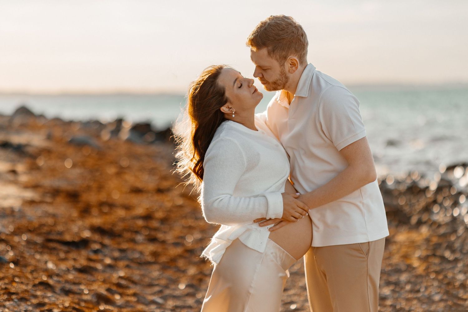 Expectant couple in white shirts and beige pants embrace tenderly on a rocky beach at golden hour, with the ocean glowing behind them.