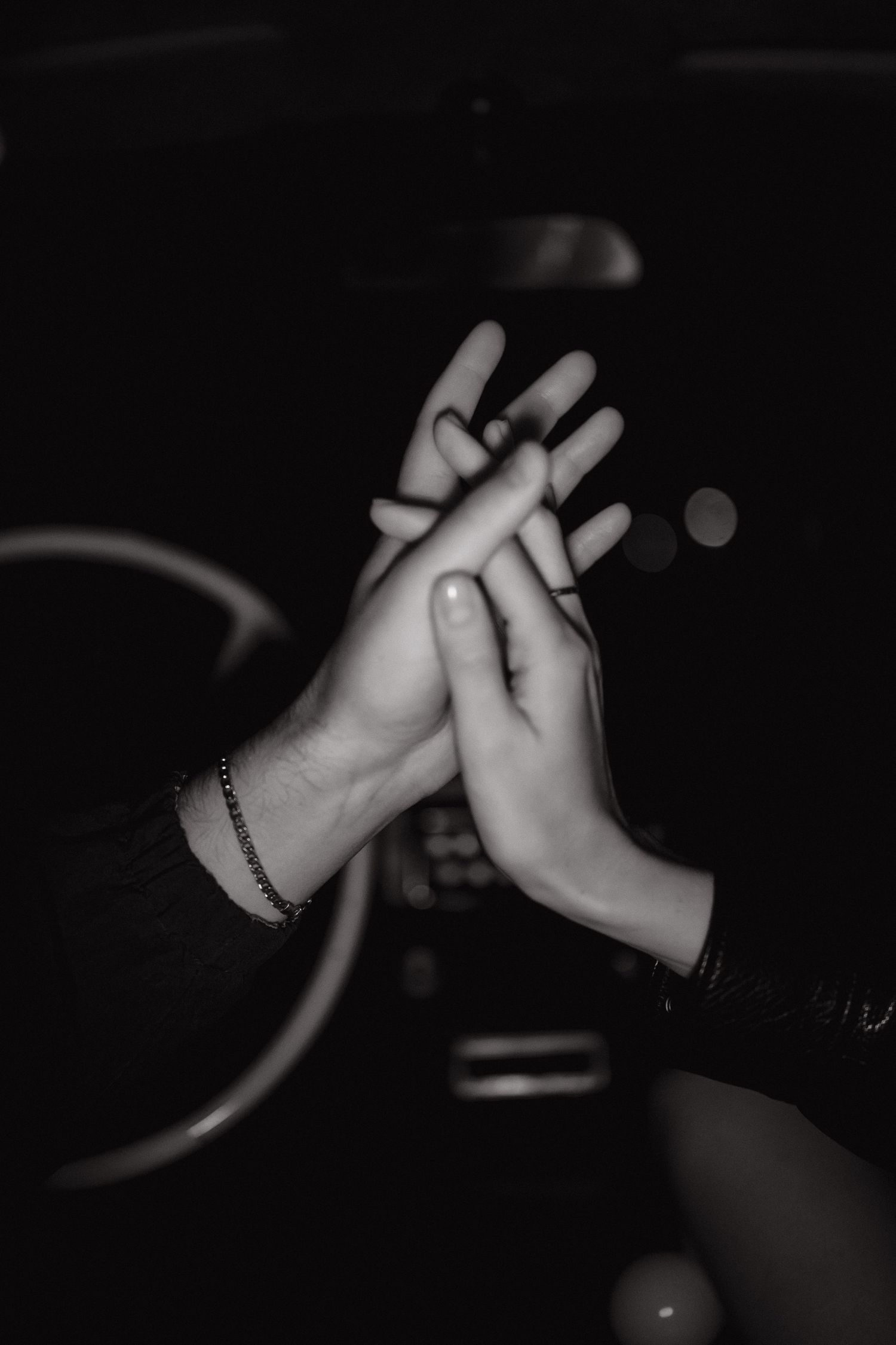 Black and white close-up of two hands touching with fingers intertwined in a dark car interior, steering wheel softly visible behind.