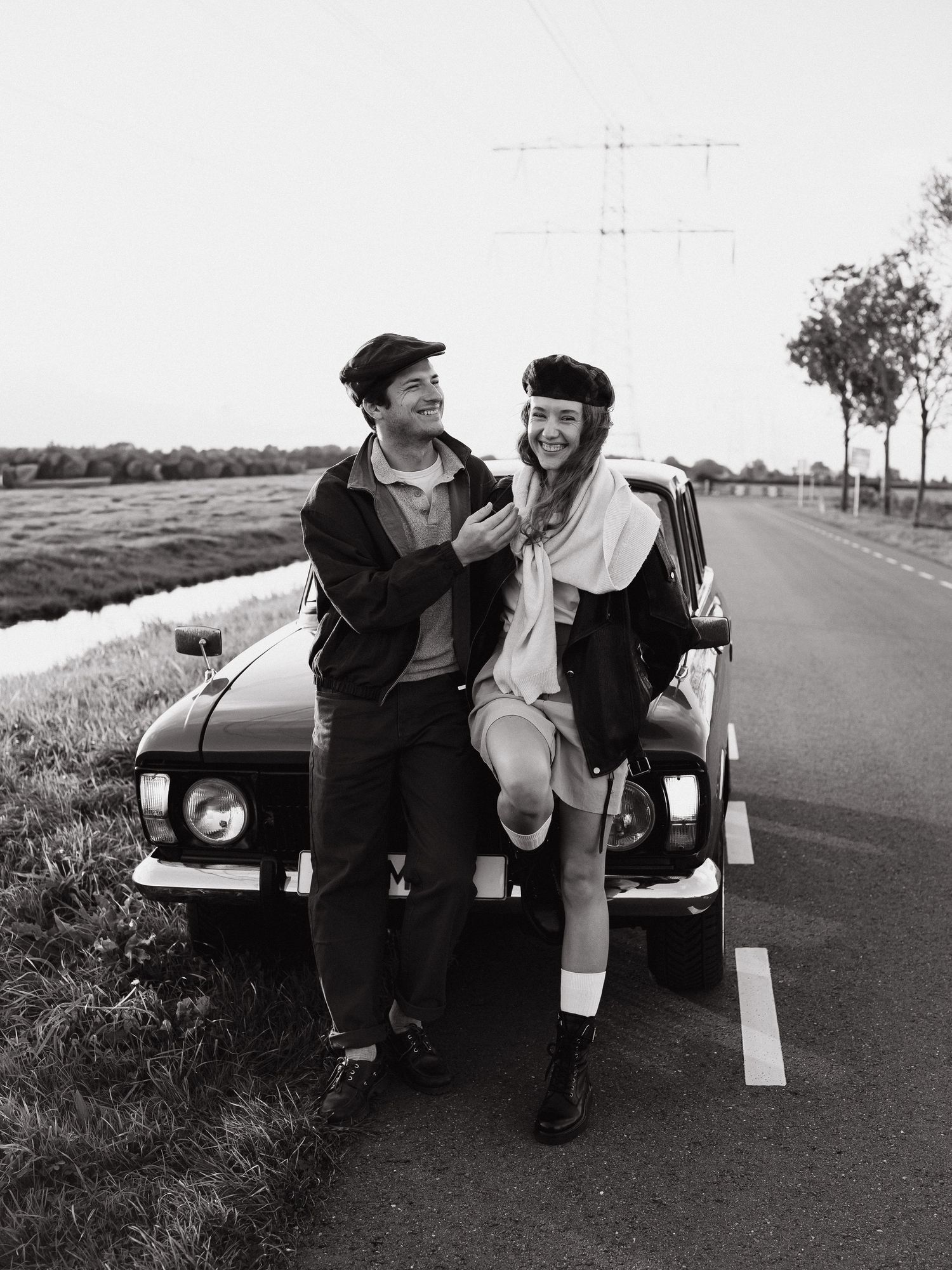 Couple sitting on the hood of a vintage car beside a country road