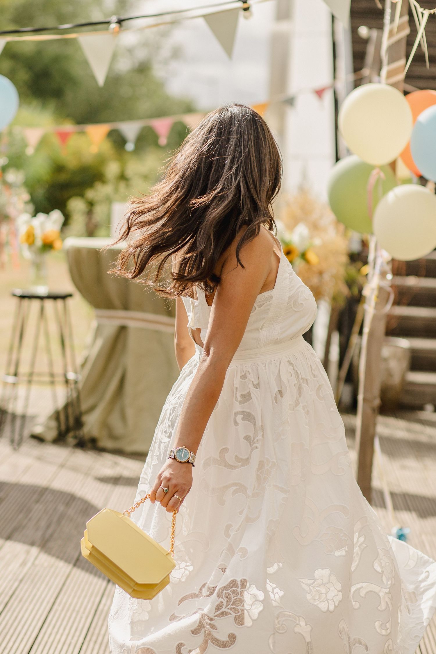 Woman in a white floral lace dress holding a yellow handbag at an outdoor party with balloons, bunting and flowers.