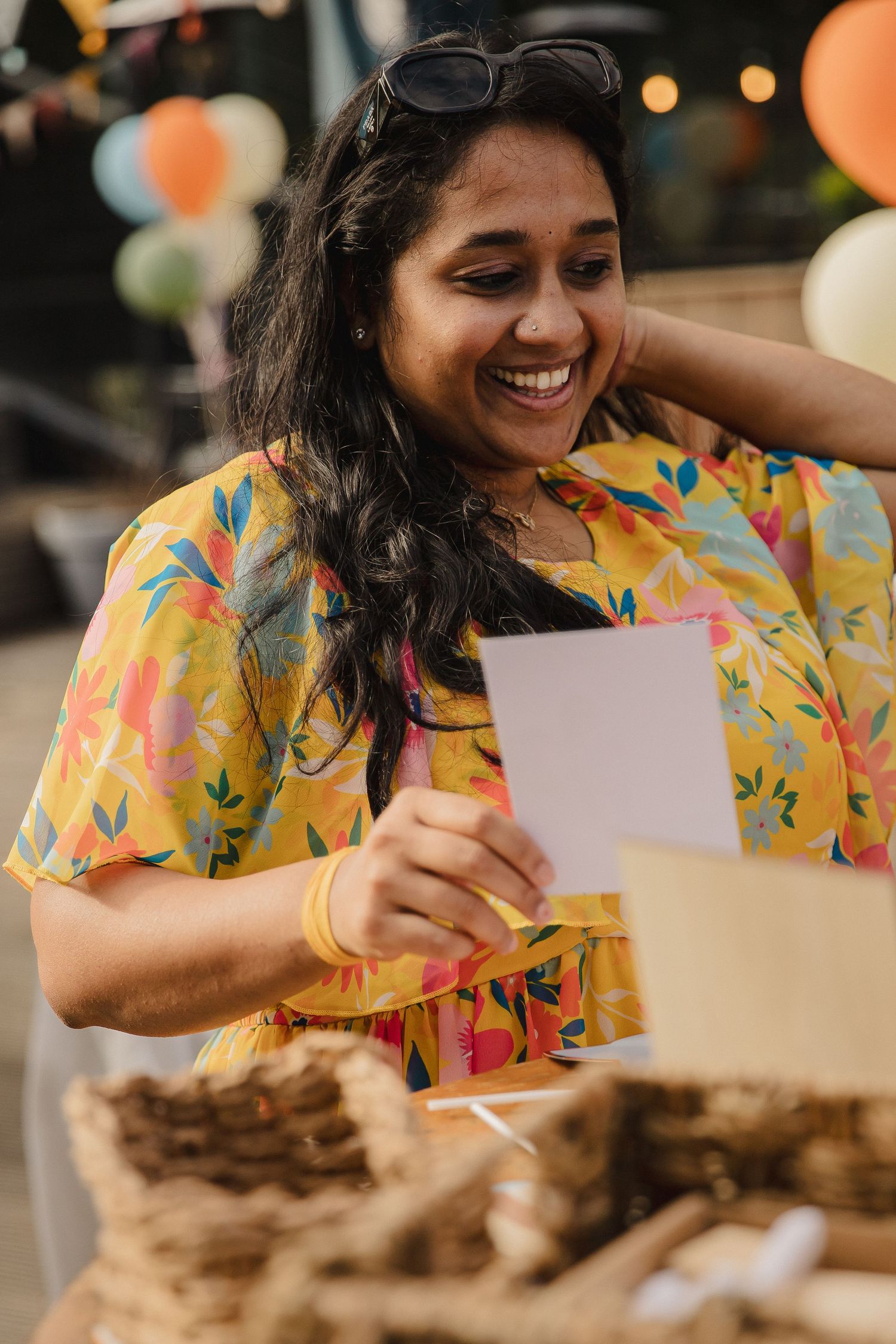 Smiling woman in a colorful floral dress holds a paper card at an outdoor party with balloons and wicker baskets nearby.