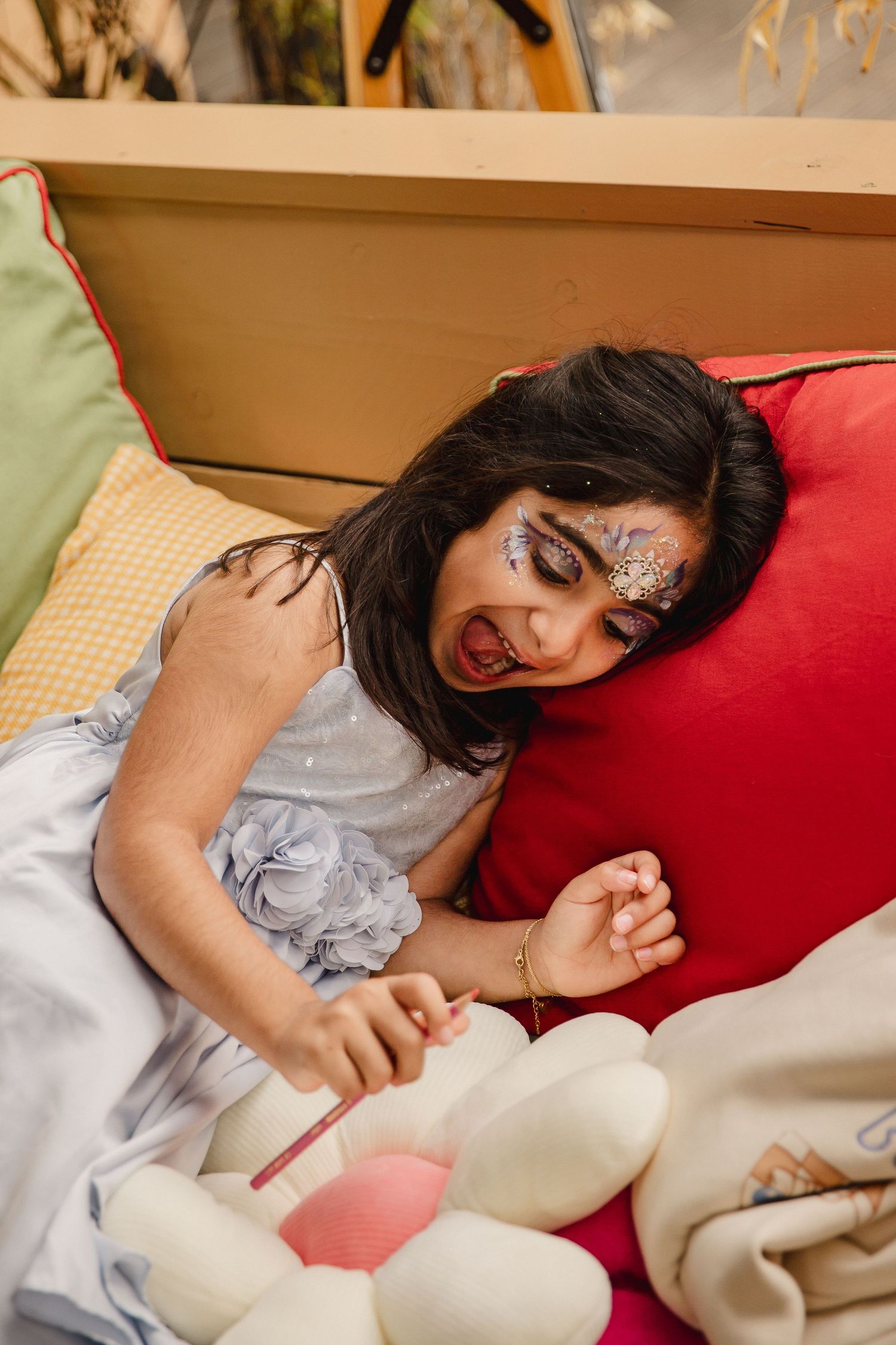 Young girl with glittery face paint in a gray dress laughs while lying on red and green cushions, holding a small brush over a plush toy.