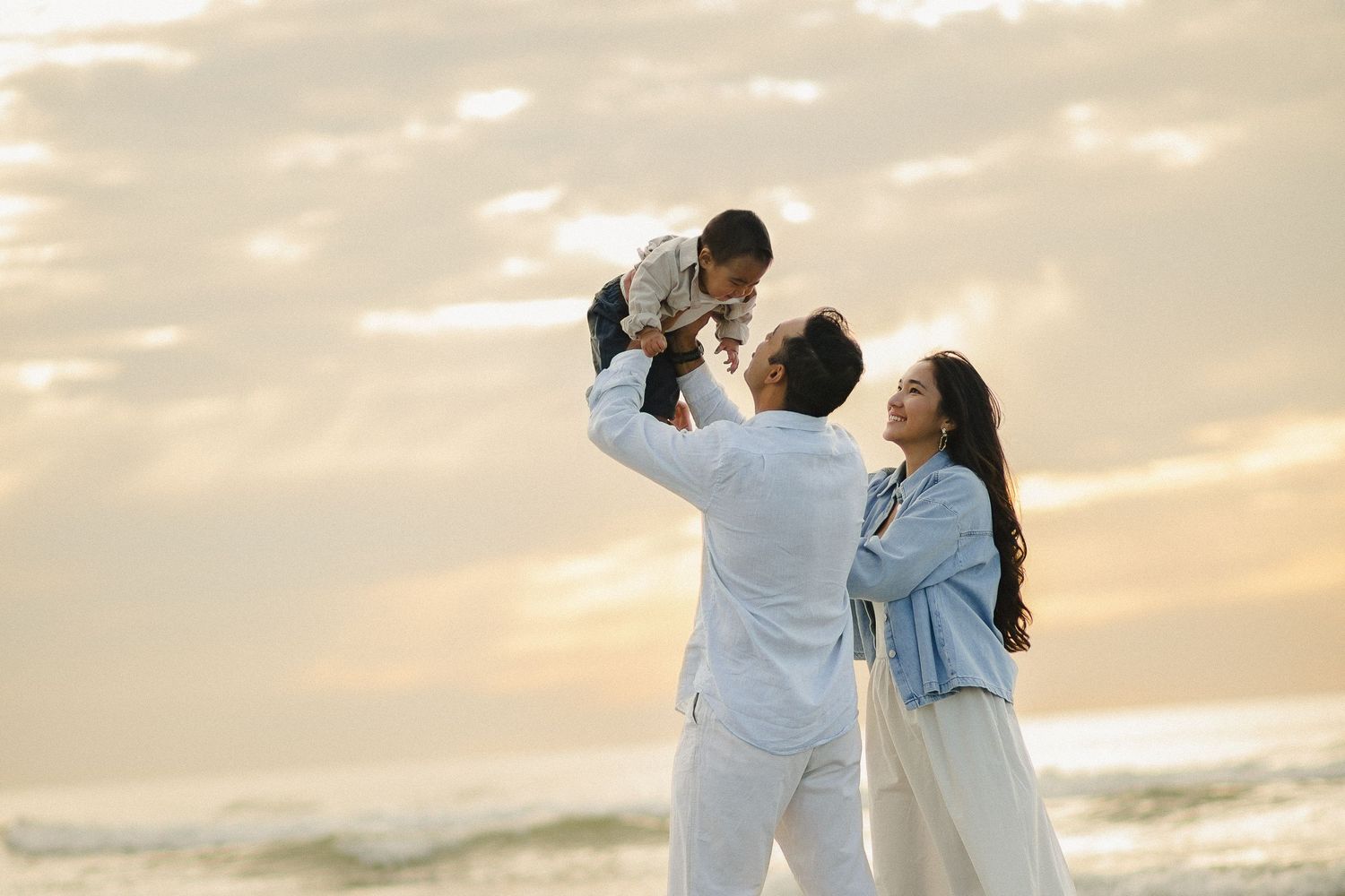 Parents lifting their baby together by the sea at sunset