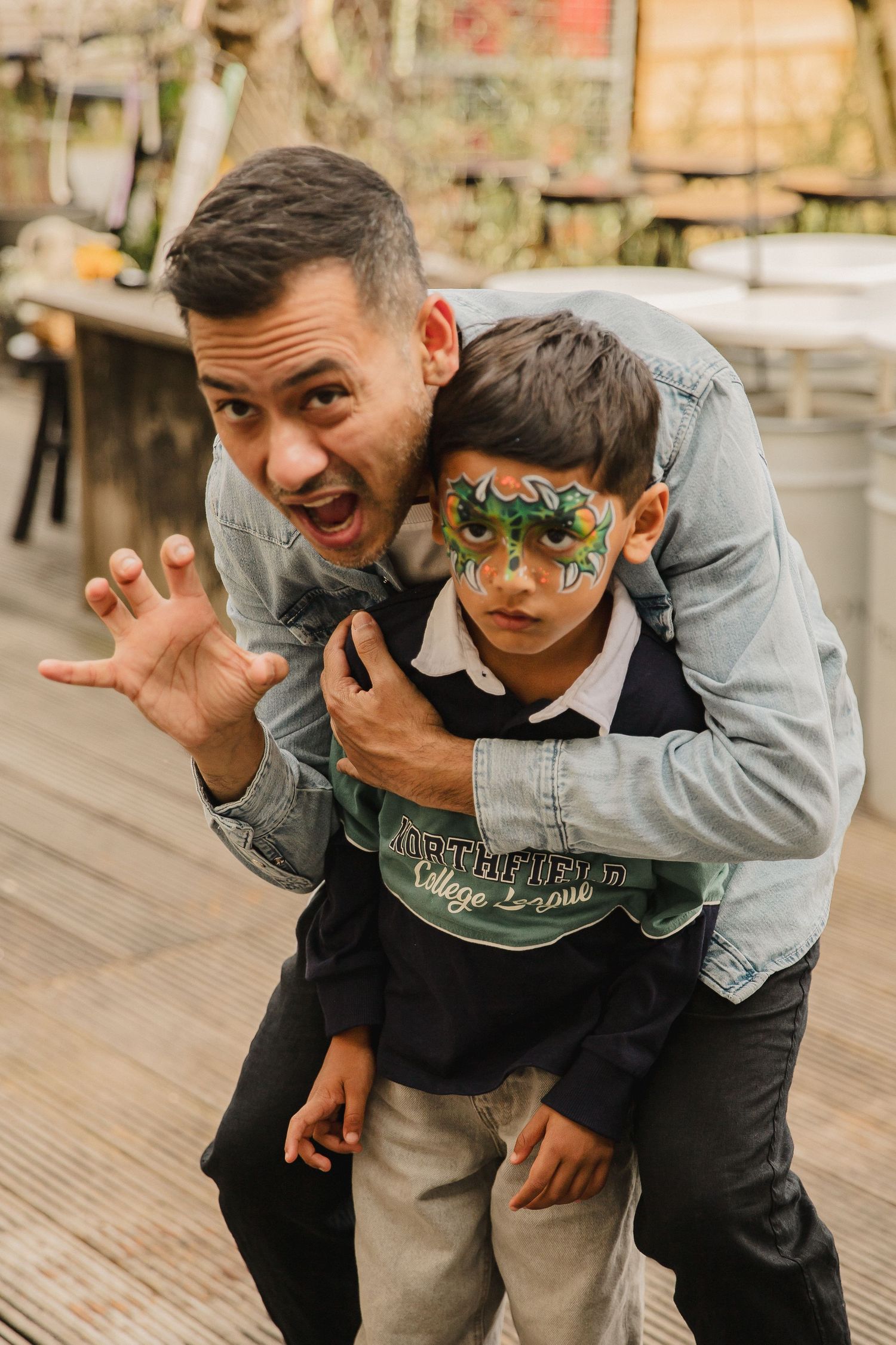 A playful man in a denim jacket makes a claw gesture while hugging a smiling boy with green face paint outdoors.
