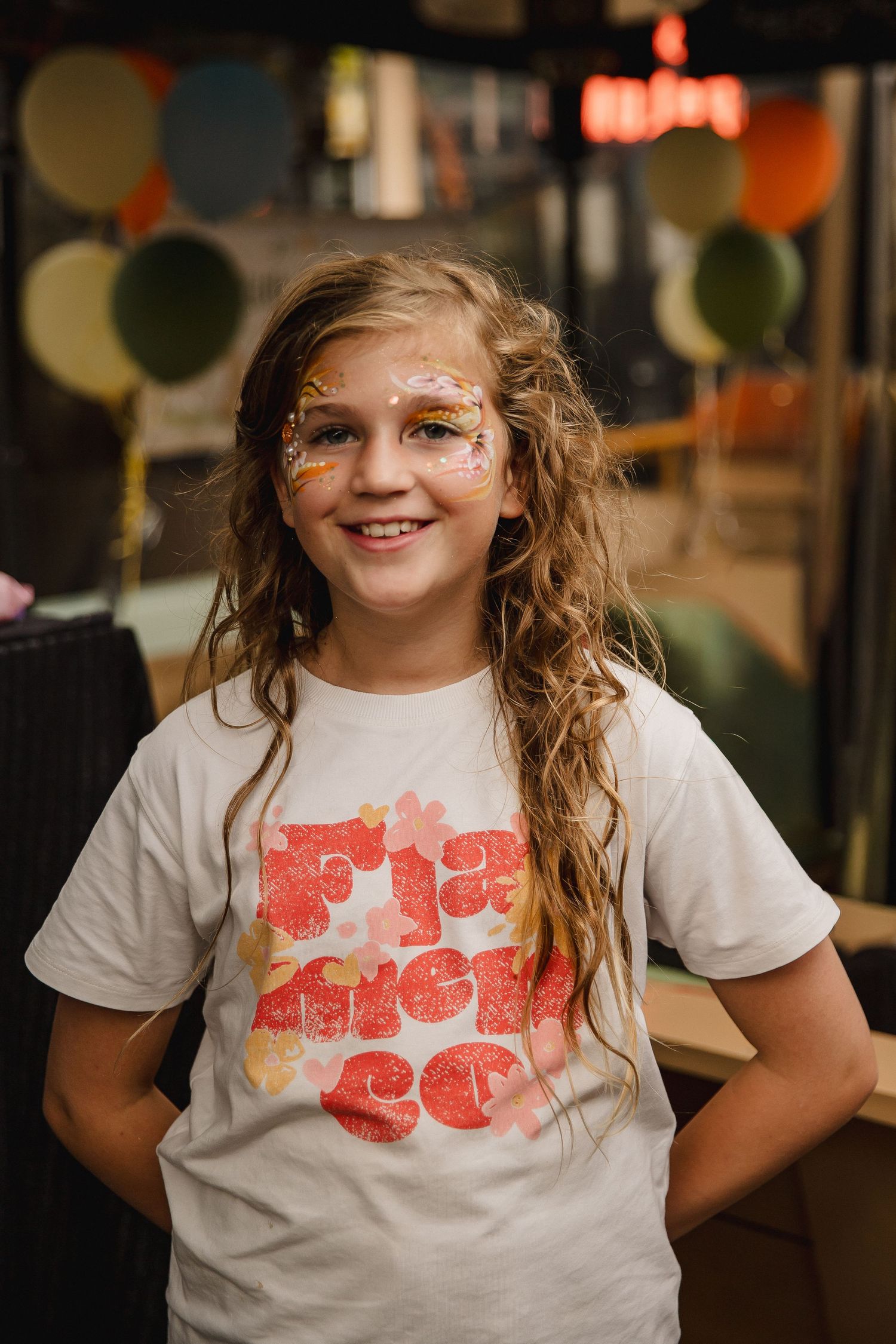 Smiling girl with long wavy hair and glittery face paint wearing a floral graphic tee at a party with balloons in the background.