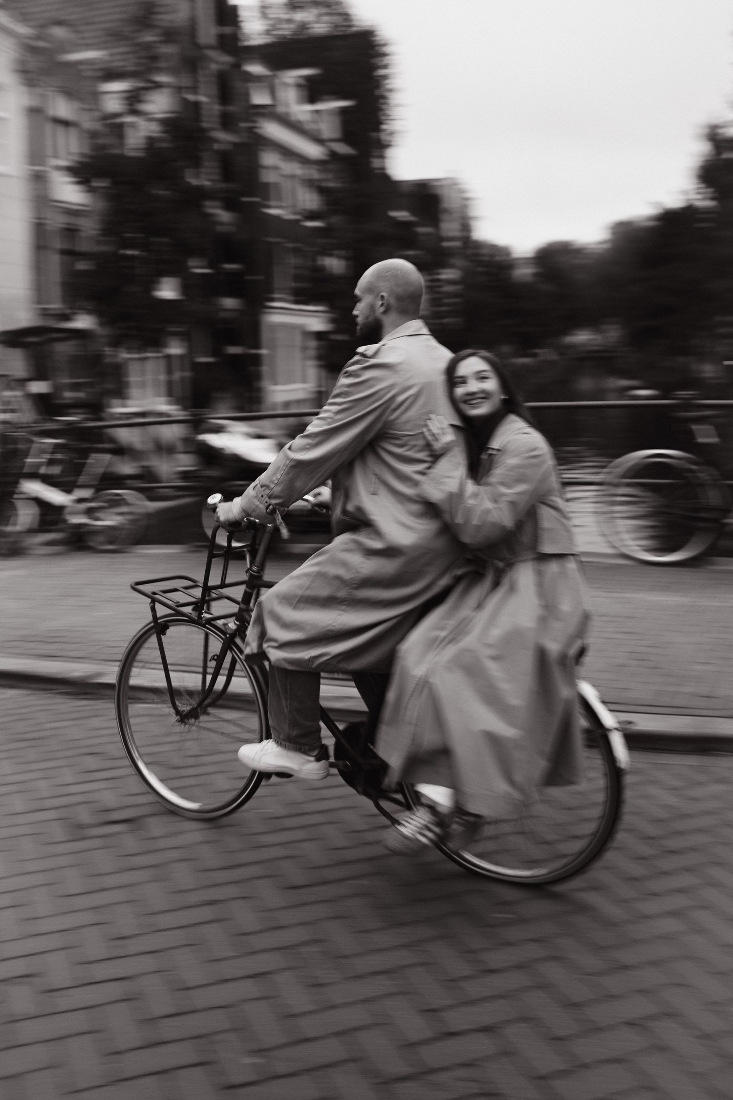Black and white motion-blur photo of a couple riding a bicycle together on a cobblestone street in Amsterdam, the woman hugging the man.
