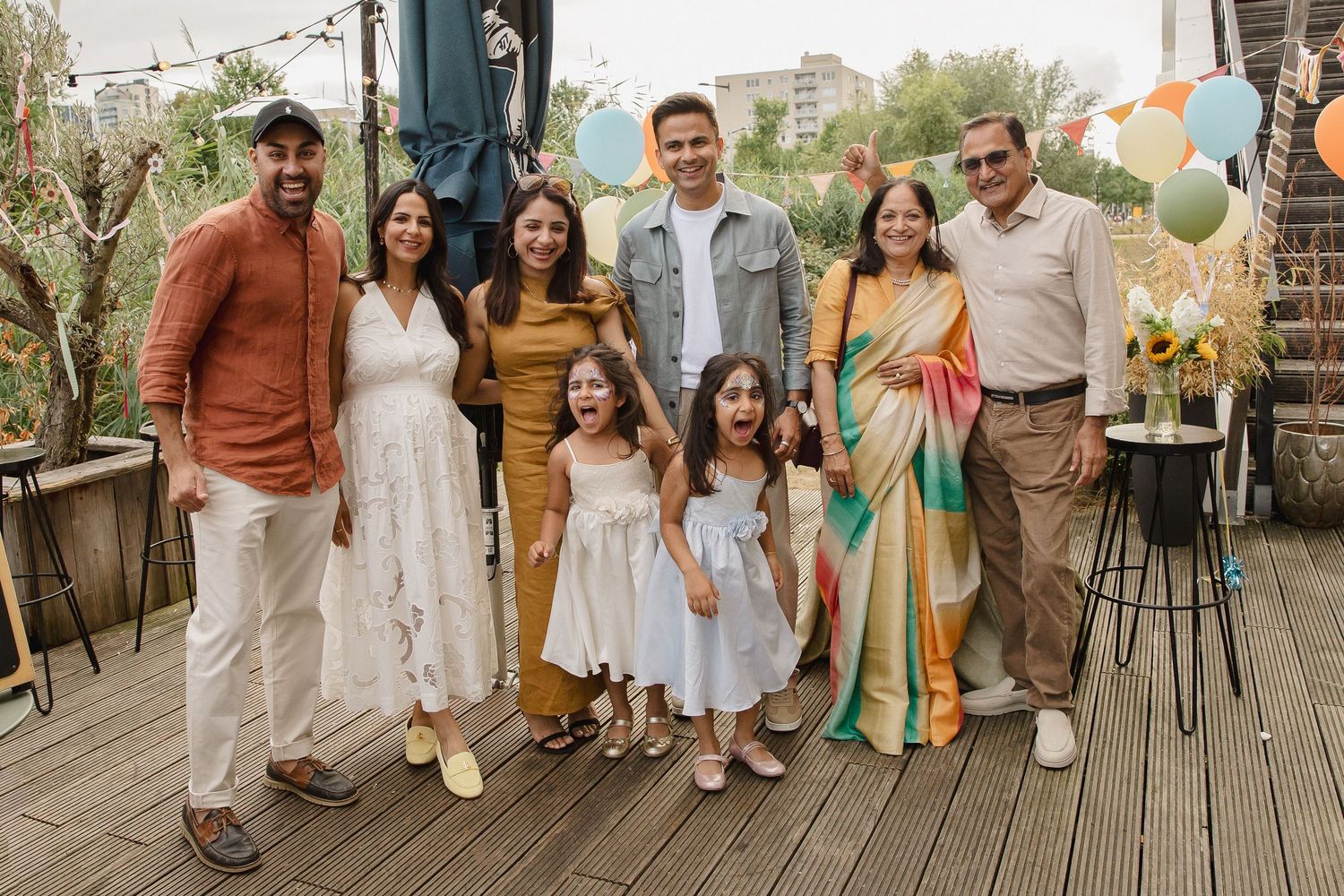 Multigenerational family of seven poses together on a sunny rooftop terrace decorated with colorful balloons during a festive celebration.