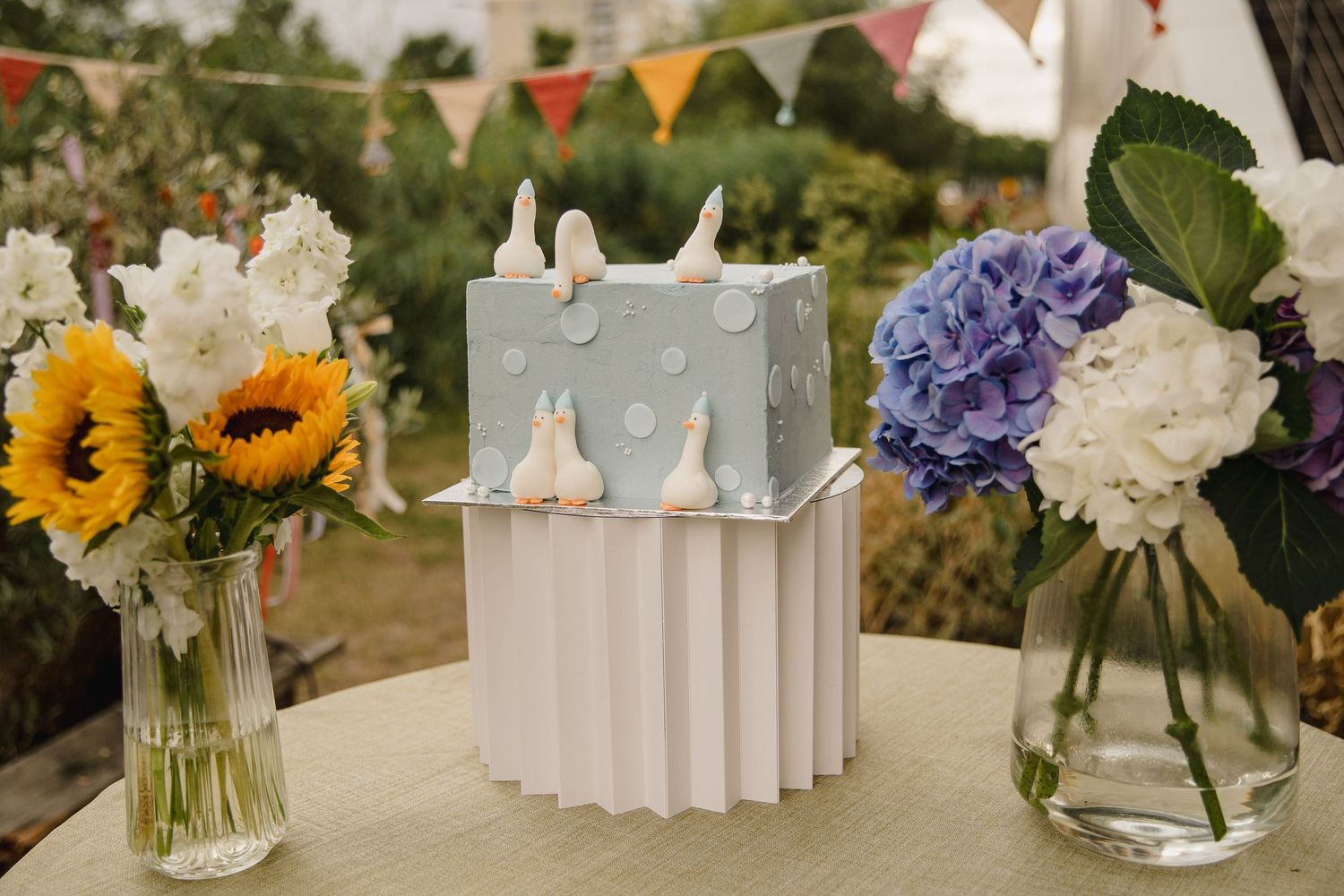 Light blue wedding cake with white bird figurines on a pleated white stand, flanked by sunflowers and hydrangeas in glass vases outdoors.