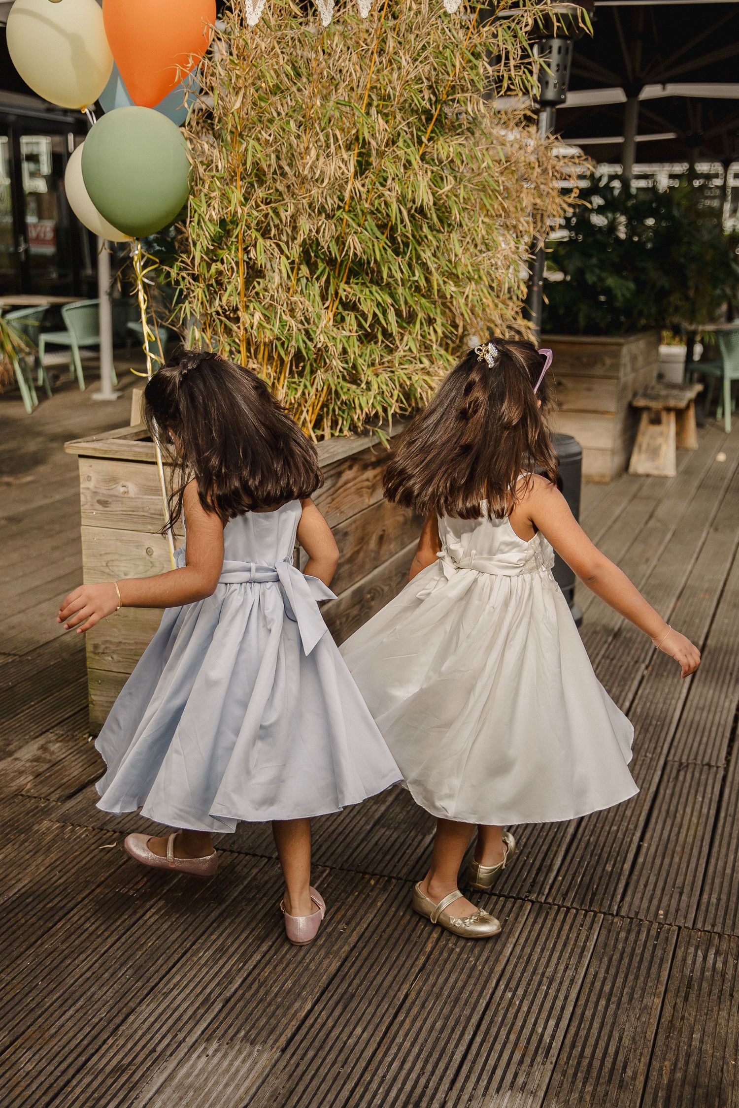Two little girls in light blue and white party dresses twirl on a wooden deck beside pastel balloons and a tall planter.